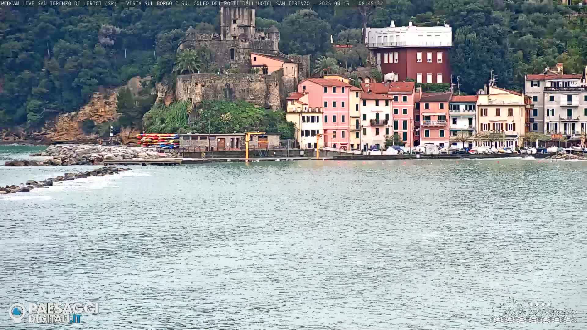 A calm body of water sits in front of a coastal town, featuring colorful buildings, a stone structure resembling a castle, and a small boat dock with kayaks.