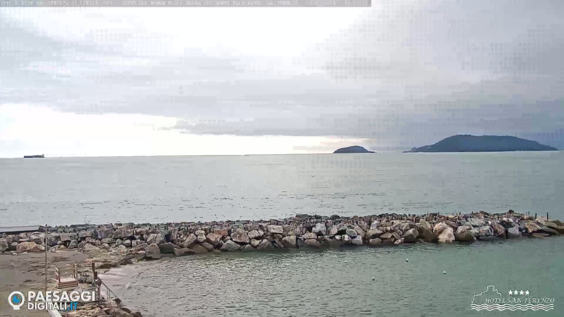 A rocky seawall separates a calm, gray sea from a sandy beach under an overcast sky, with an island visible in the distance.
