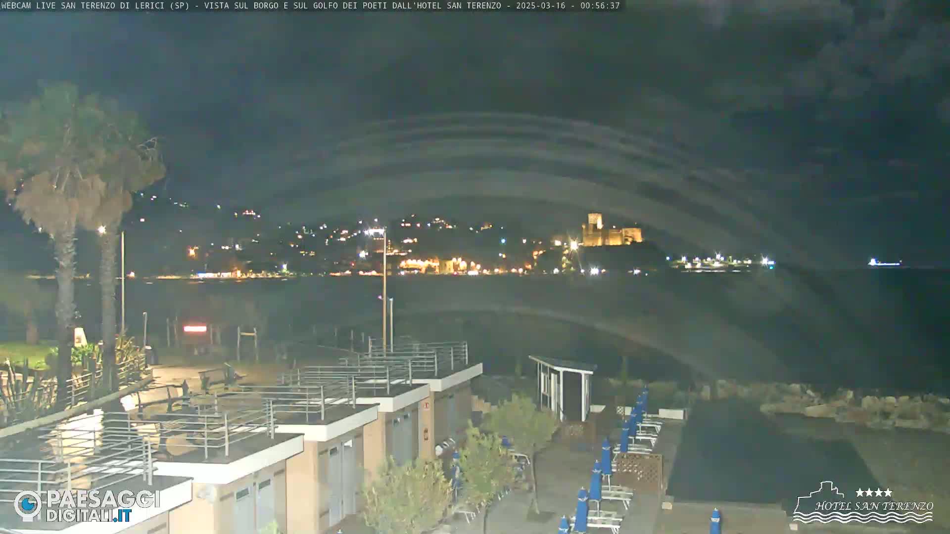 A nighttime view from a seaside resort shows a calm body of water, a distant town with lights, and a castle perched on a hill, under a cloudy sky.