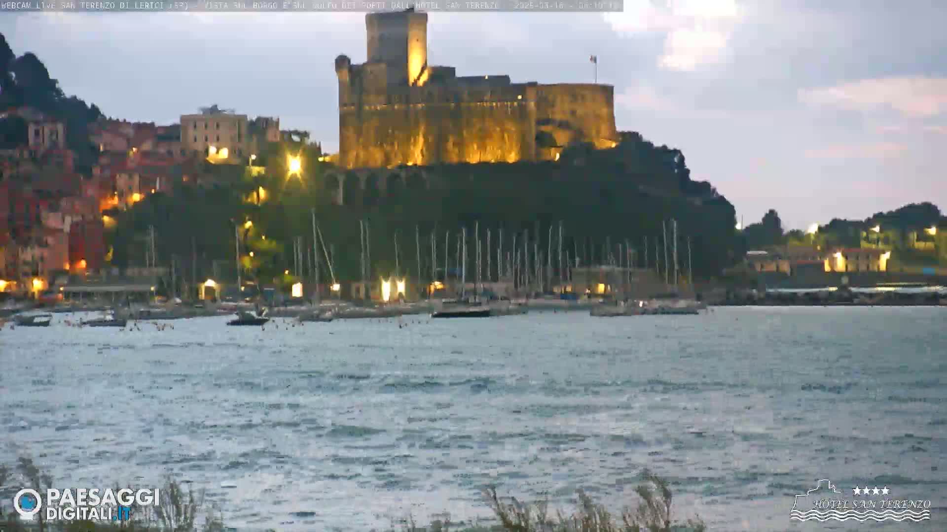 A large, illuminated castle sits atop a hill overlooking a harbor filled with boats at dusk, under a partly cloudy sky.