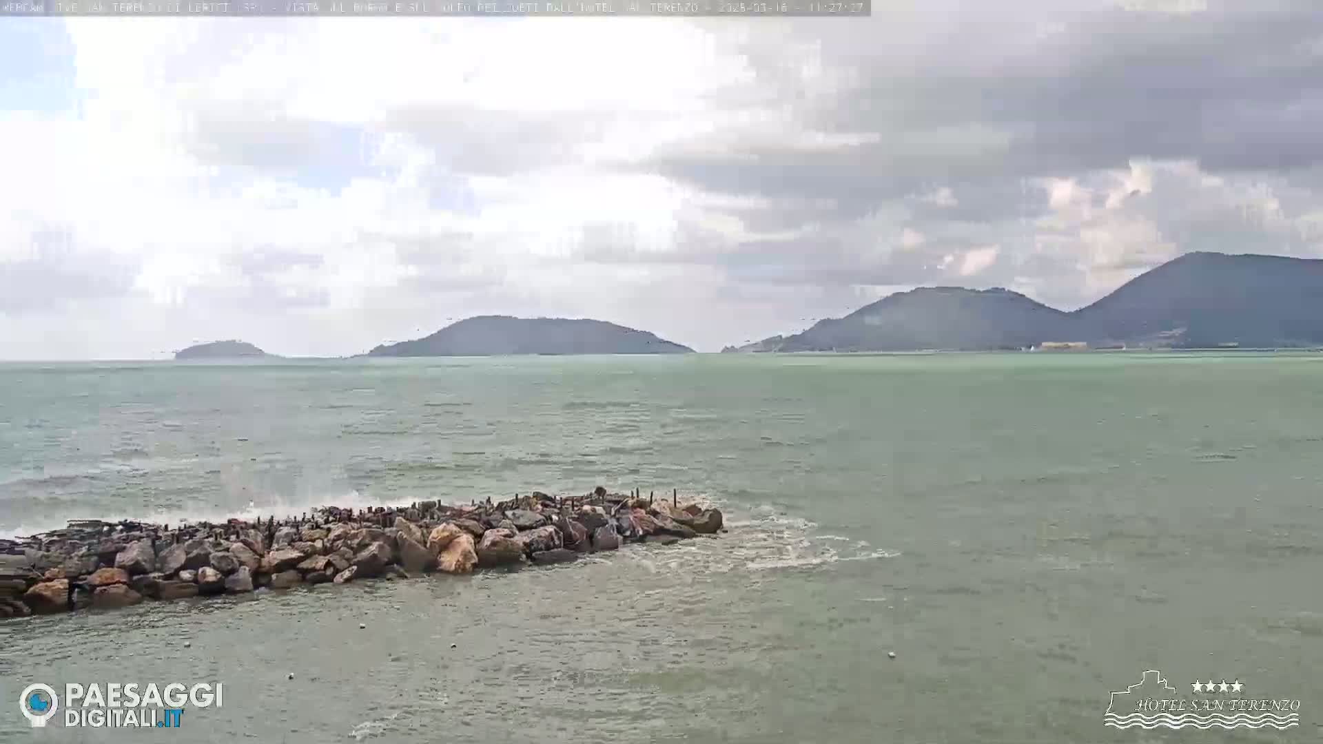 A rocky breakwater partially submerges in a somewhat choppy sea under a cloudy sky, with distant hills visible on the horizon.