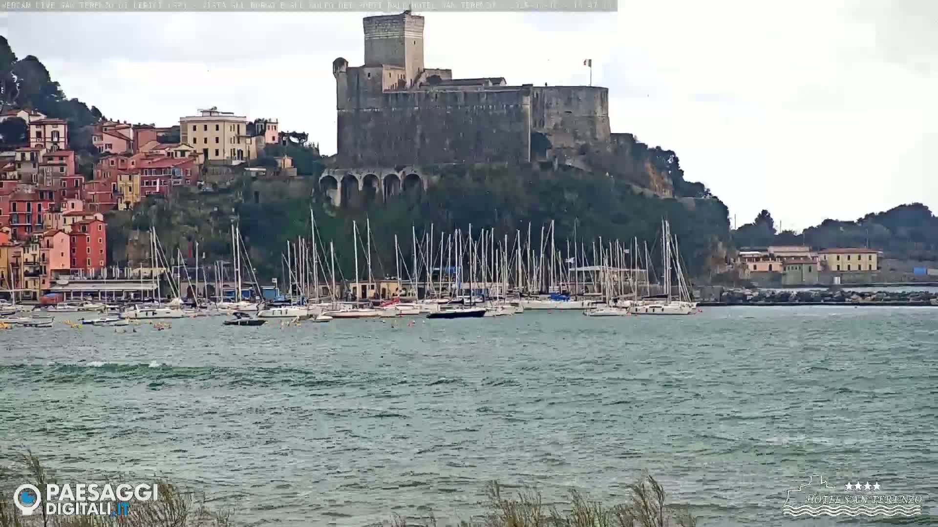 A stone castle sits atop a hill overlooking a harbor filled with sailboats on a slightly choppy, overcast day.