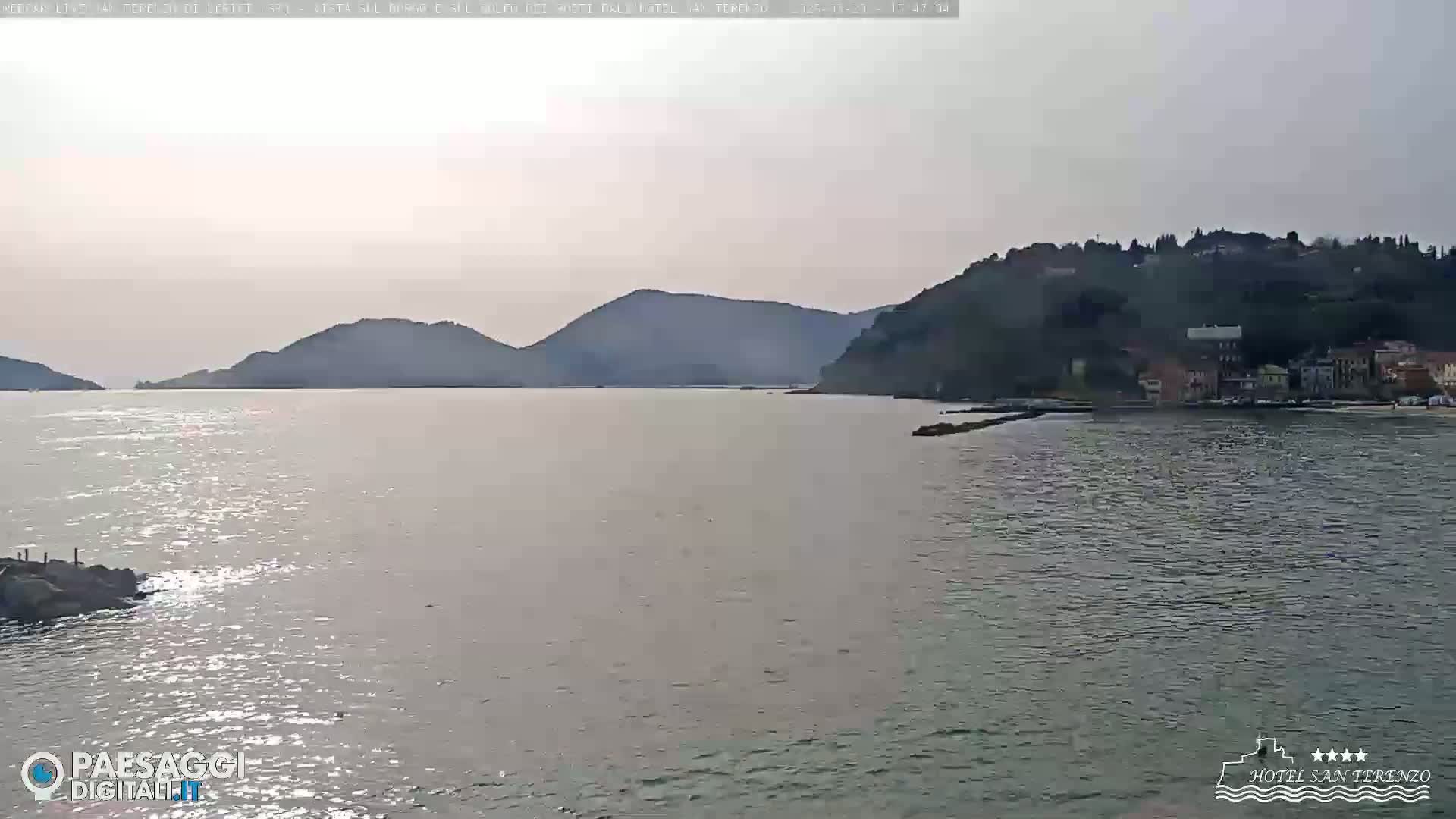 A calm body of water stretches out to meet hazy hills and a small village under a pale, overcast sky.