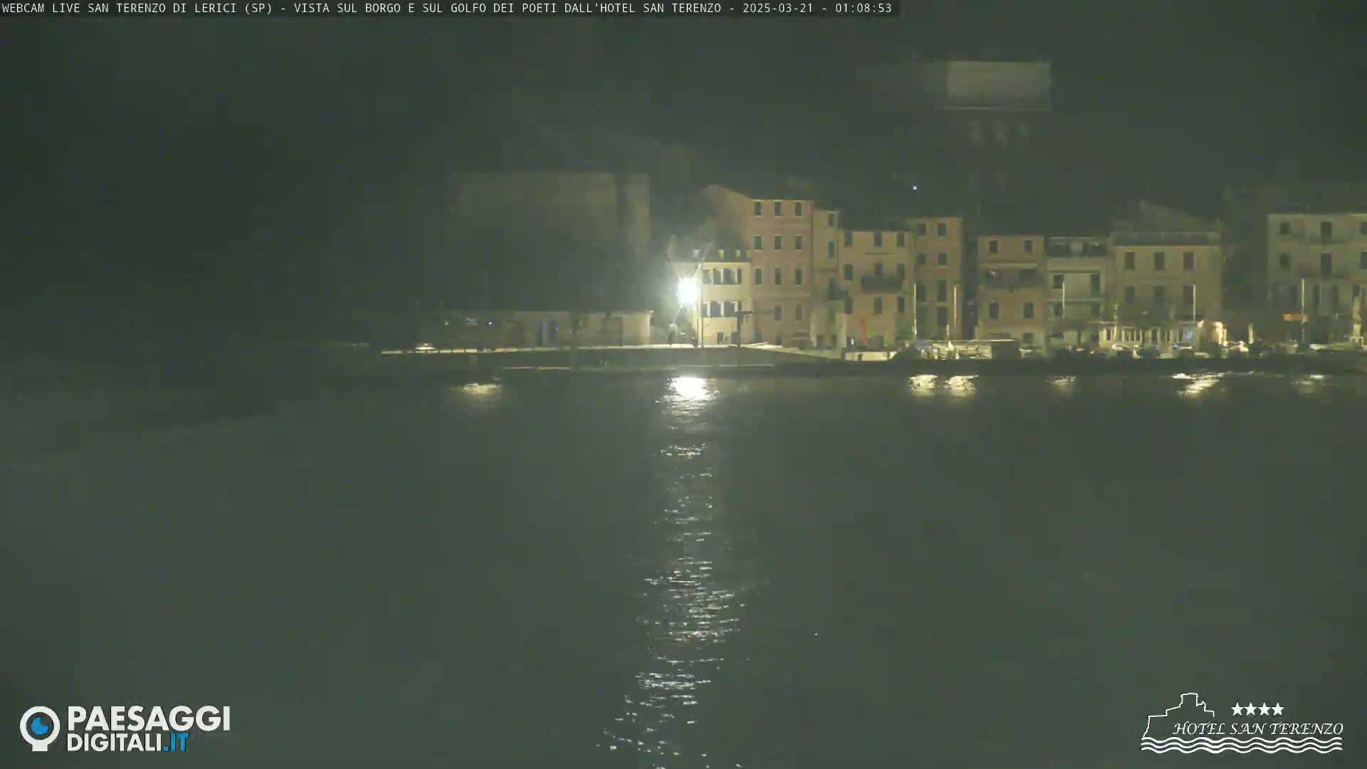 A nighttime view across calm water shows a row of illuminated buildings along a waterfront.
