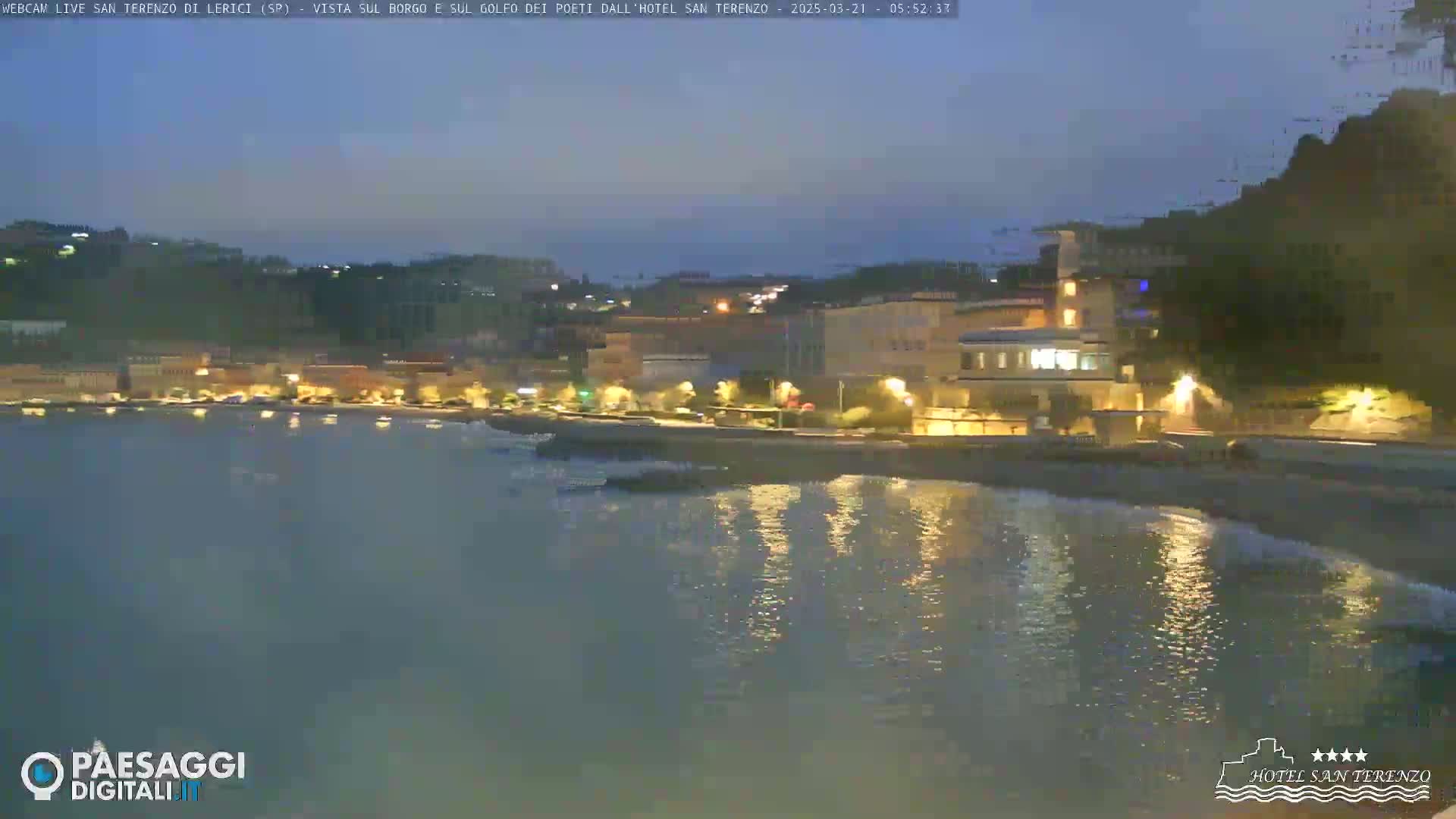 A nighttime view of a seaside town with lights reflecting on calm water under a dark sky.
