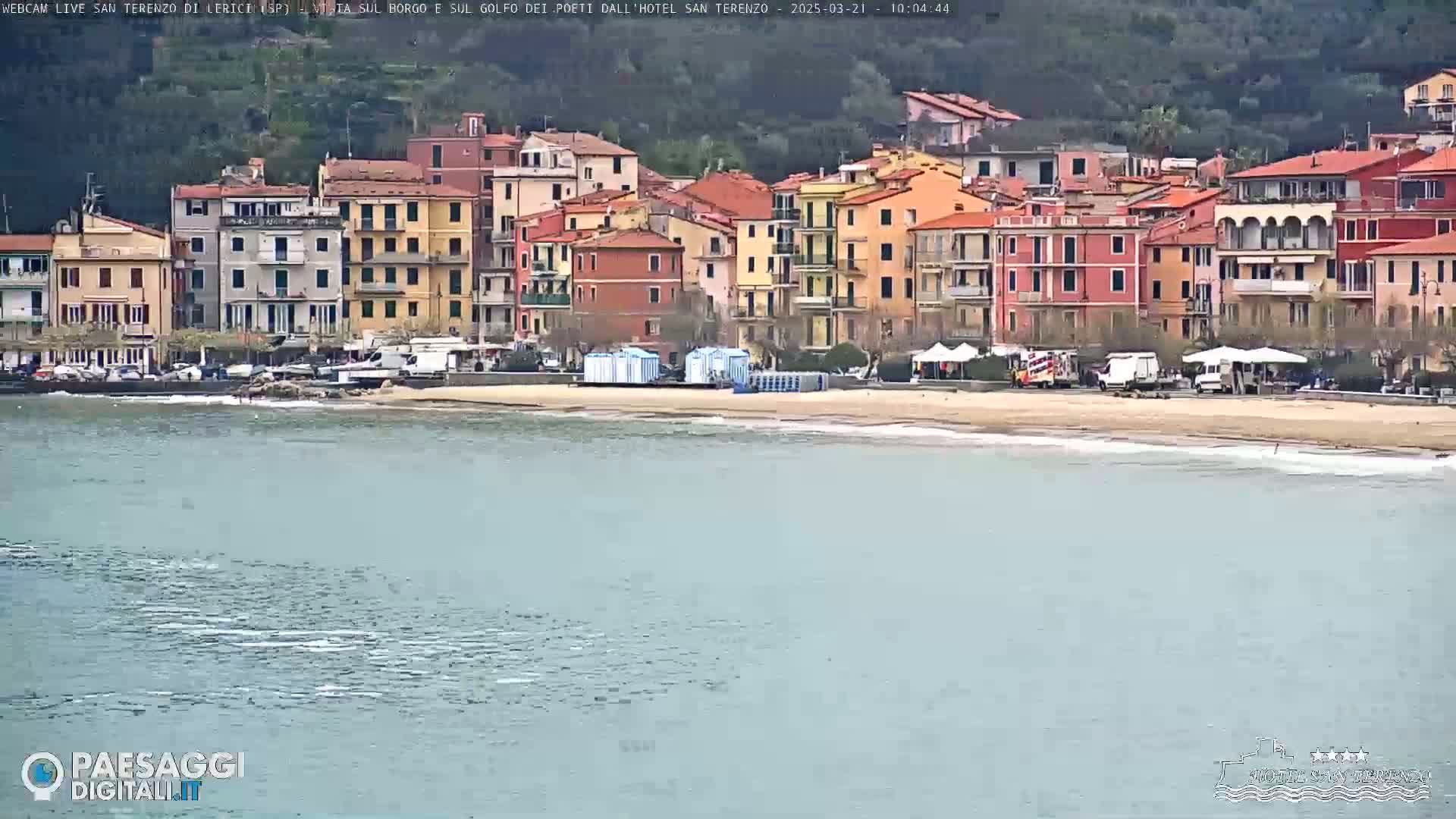 A seaside town with colorful buildings sits on a sandy beach next to calm, light-blue water under an overcast sky.