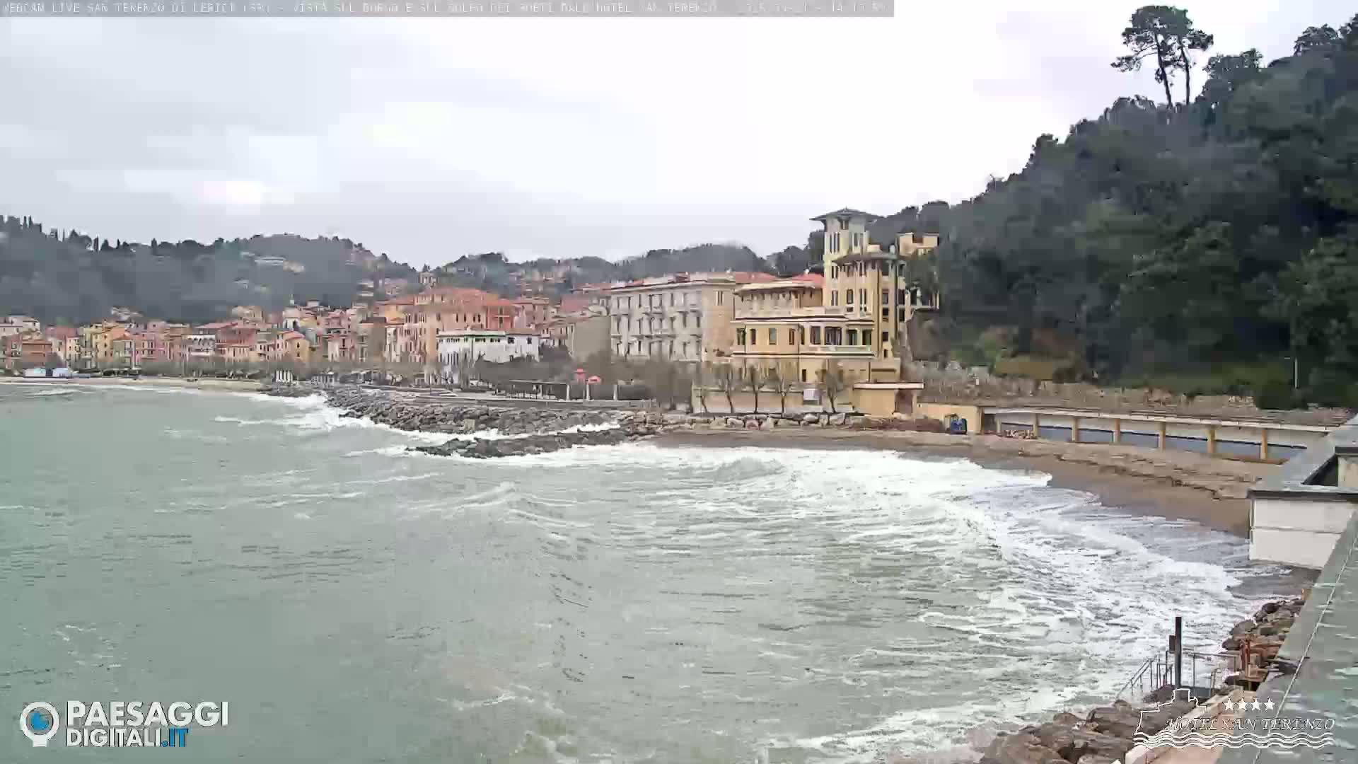 Waves crash on a sandy beach in front of a seaside town nestled against a hillside under an overcast sky.