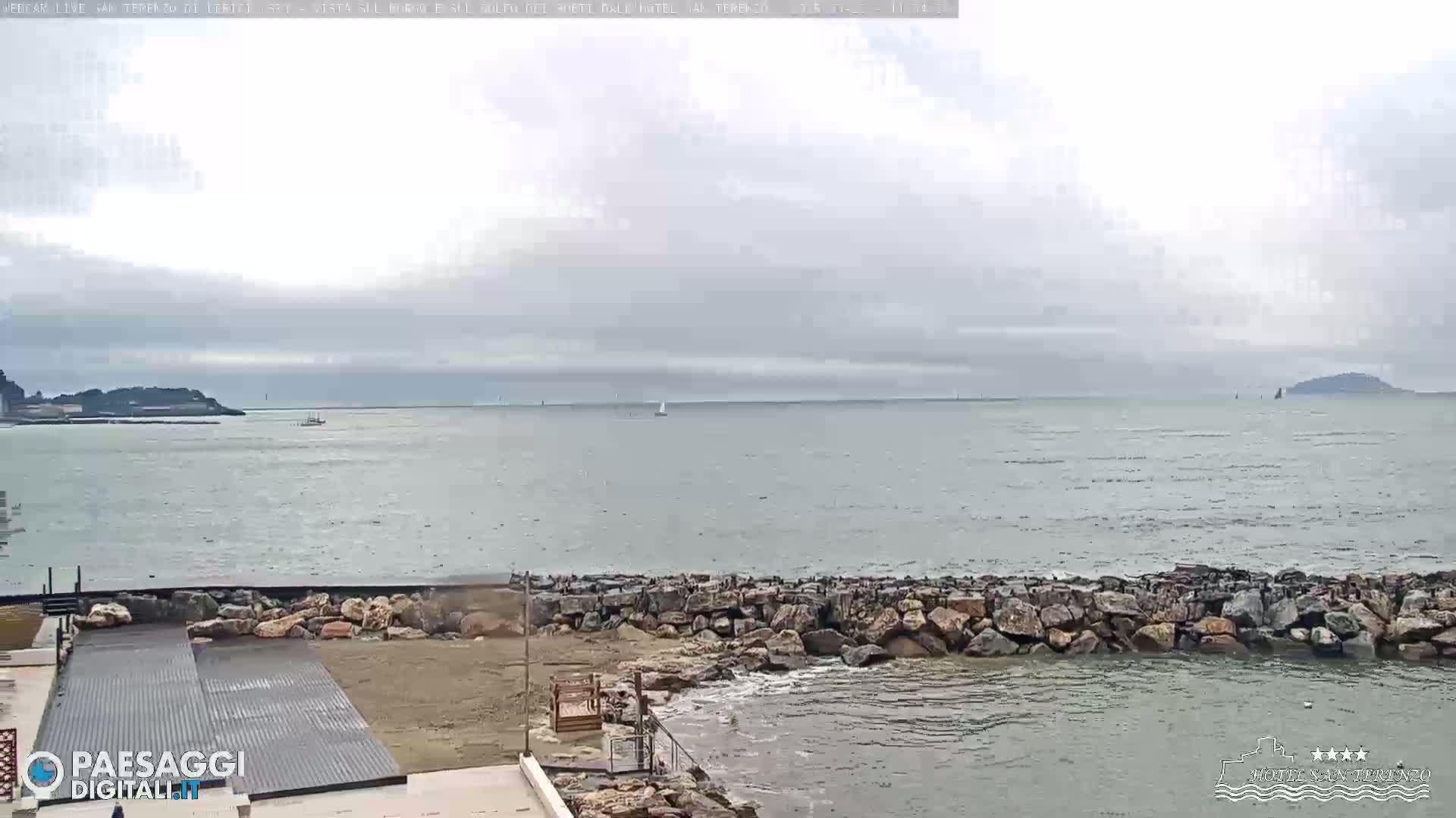 A rocky shoreline and calm, grey sea stretch to distant land under an overcast sky.