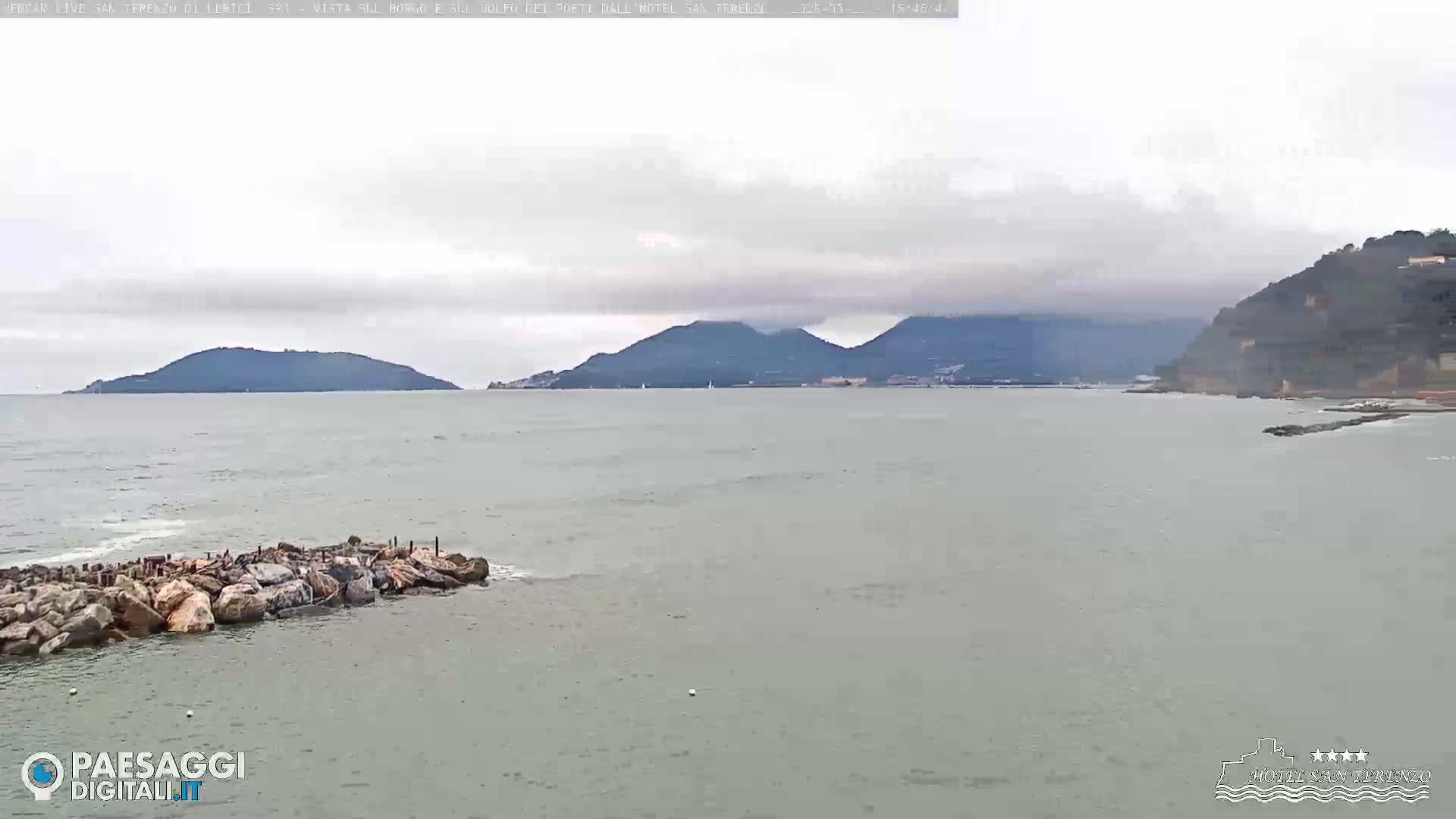 A calm sea with a rocky outcrop in the foreground, overlooking distant hills and mountains under an overcast sky.