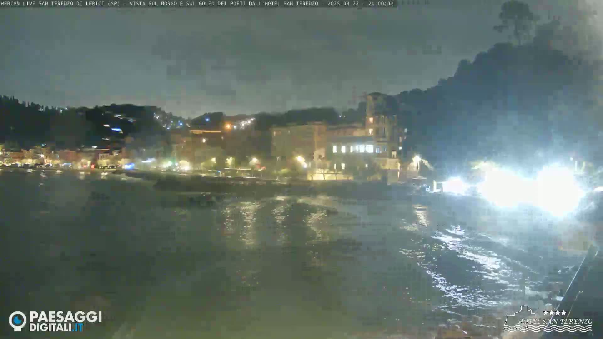 A nighttime coastal town is seen with choppy water under a dark, overcast sky.