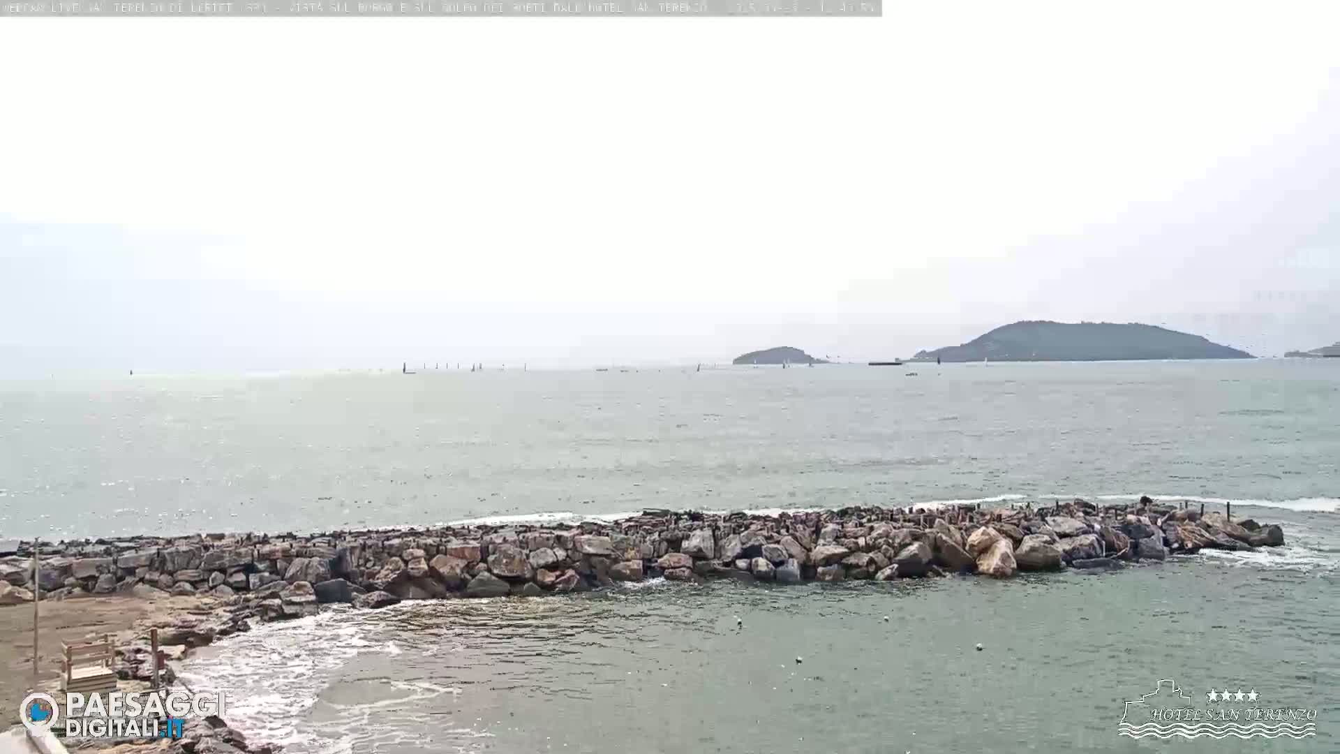A rocky breakwater protects a calm, gray-green sea from small waves under an overcast sky, with distant islands visible on the horizon.