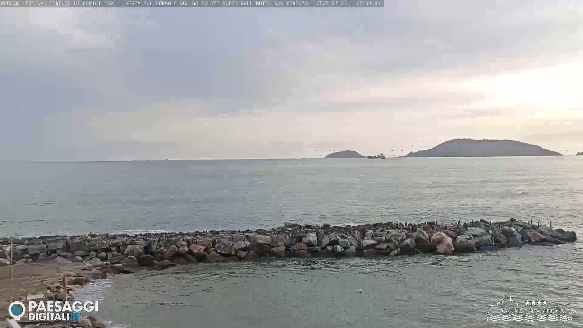 A rocky breakwater extends into a calm sea under a mostly cloudy sky, with a distant island visible on the horizon.
