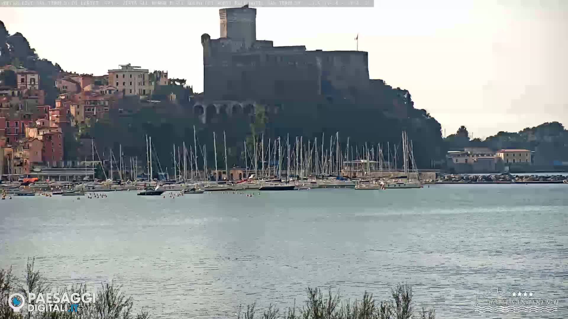 A stone castle sits atop a hill overlooking a calm body of water filled with numerous sailboats docked at a marina, with a town nestled at its base.