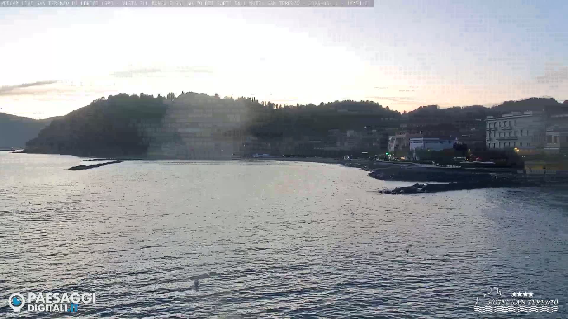 A calm body of water laps against a rocky shoreline, with a hill dotted with buildings in the background under a twilight sky.