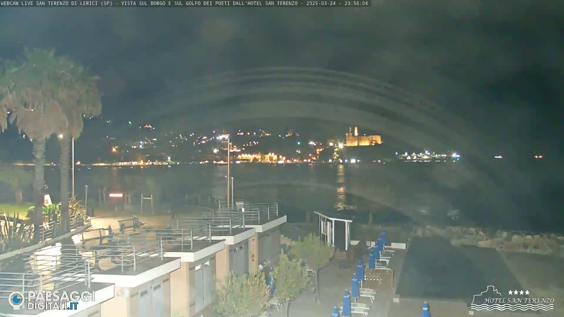 A nighttime view of a calm body of water with a town's lights visible across it, a beach with beach umbrellas and some buildings in the foreground.