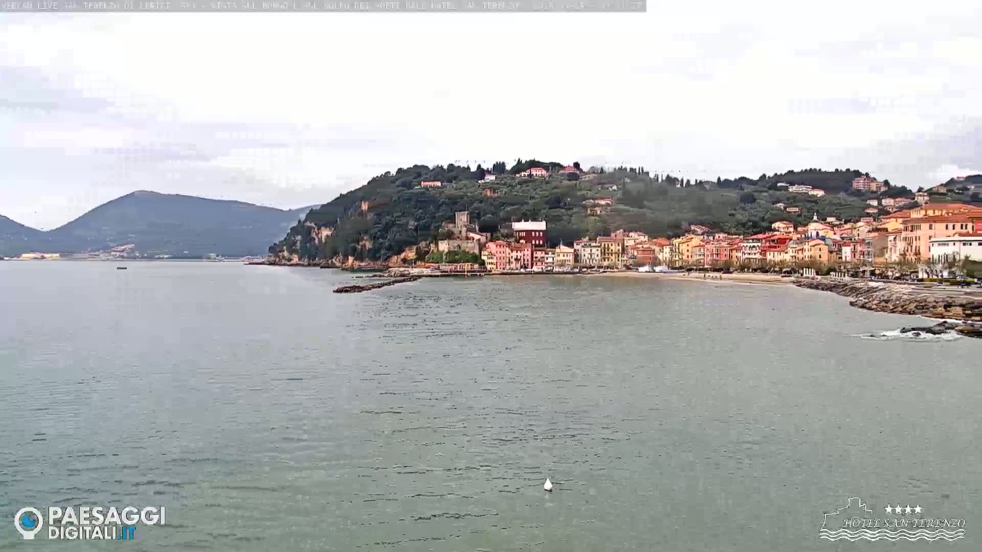 A coastal town with colorful buildings nestled against a hillside overlooks a calm, gray sea under an overcast sky.