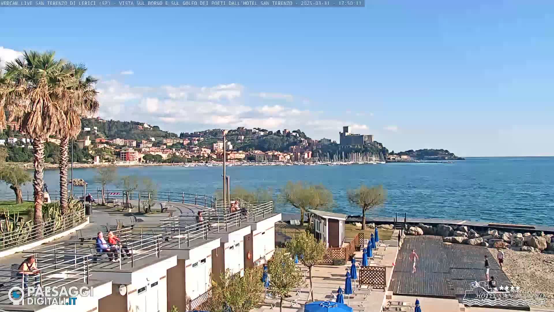 A seaside town with a castle on a hill is visible across a calm bay from a sunny beachfront area with palm trees, beach umbrellas, and a few people relaxing.
