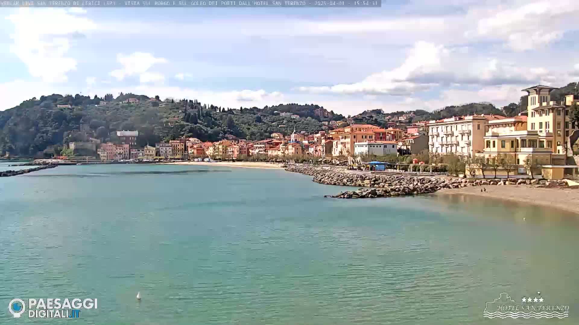 A mostly sunny day shows a seaside town nestled against a green hillside, with a calm, teal-colored body of water in the foreground.