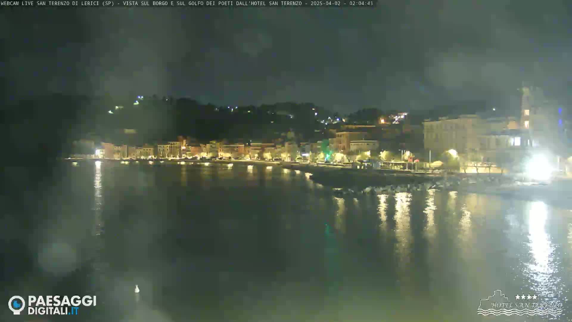 A nighttime view of a coastal town with lights reflecting on calm water under a dark, slightly hazy sky.
