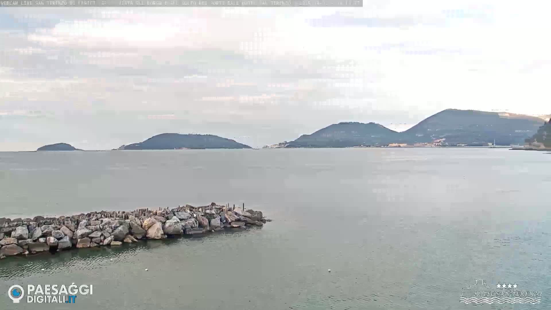 A rocky breakwater extends into a calm, gray sea, with several hills and mountains visible in the distance under a cloudy sky.