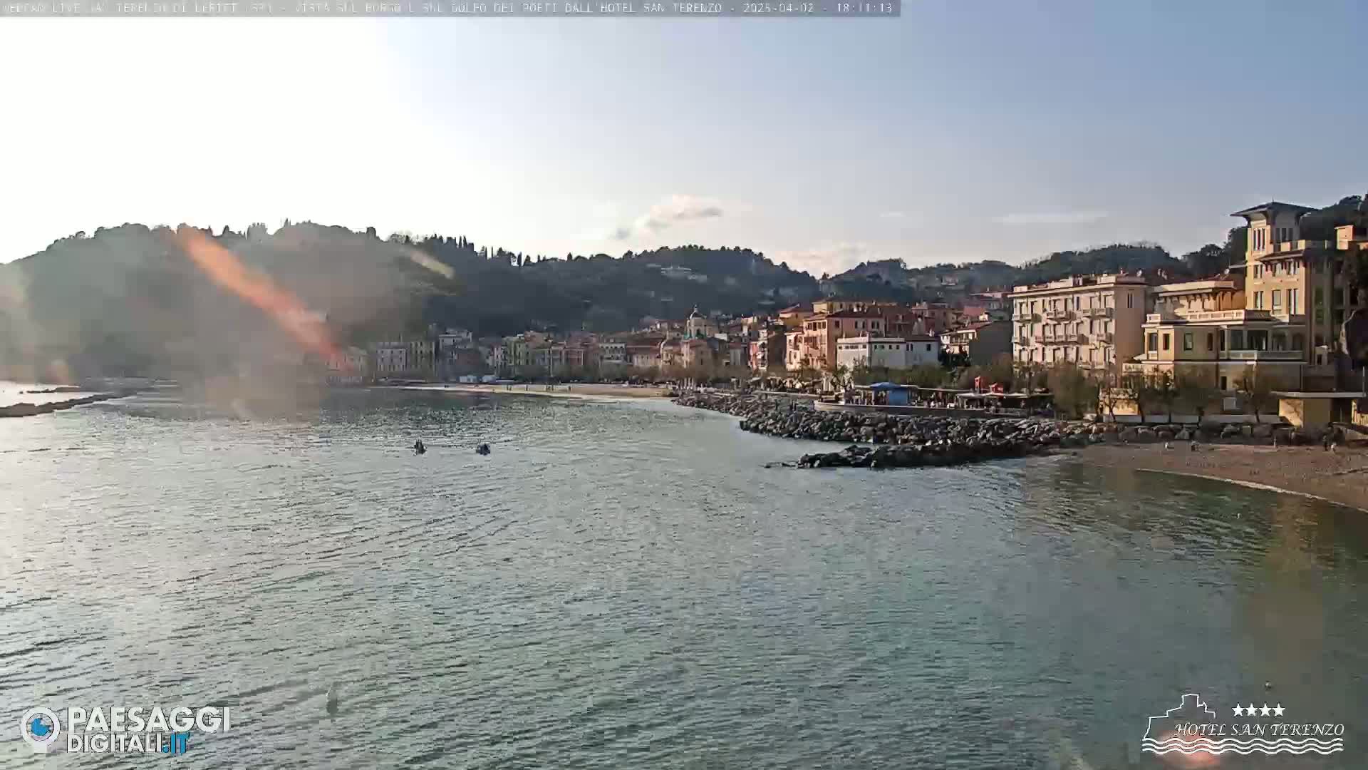 A calm body of water laps a pebble beach in front of a coastal town nestled against hills under a mostly clear sky.