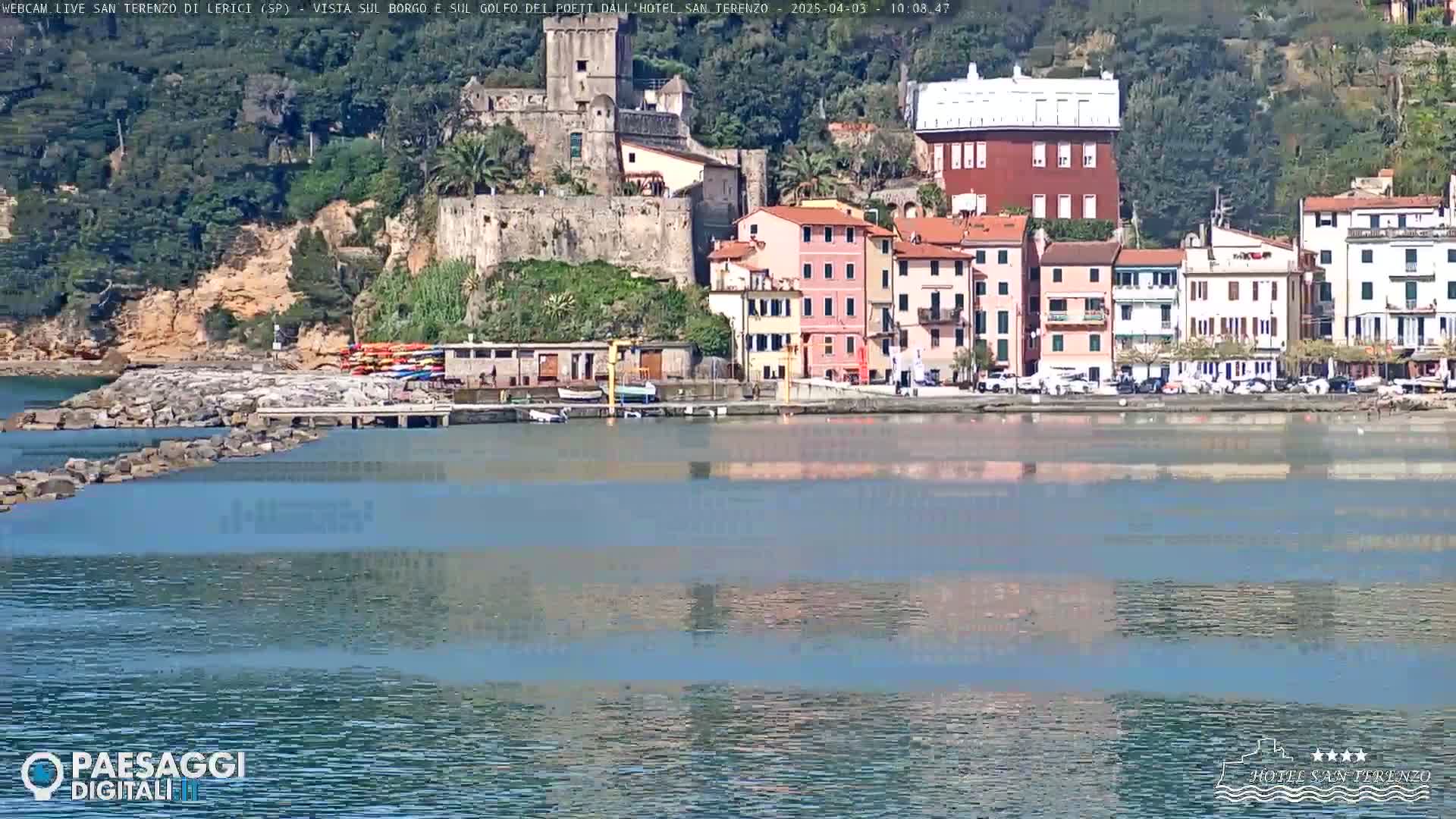 A calm body of water reflects a waterfront town nestled against a hillside under a sunny sky.