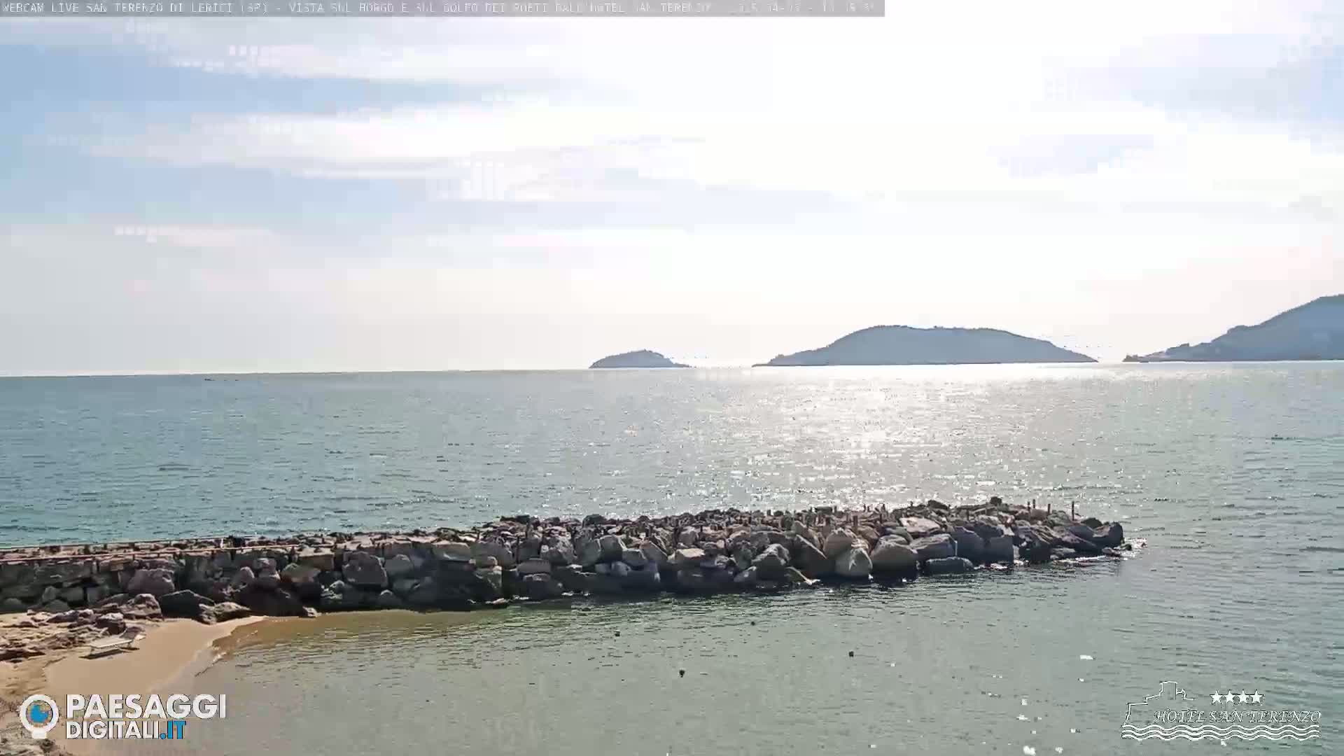 A rocky breakwater protects a small sandy beach, overlooking a calm sea with distant hills under a mostly clear sky.