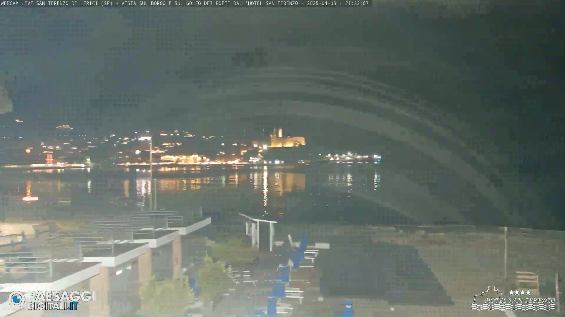 A nighttime view of a calm body of water reflecting the lights of a town across the water, with some outdoor furniture visible in the foreground.