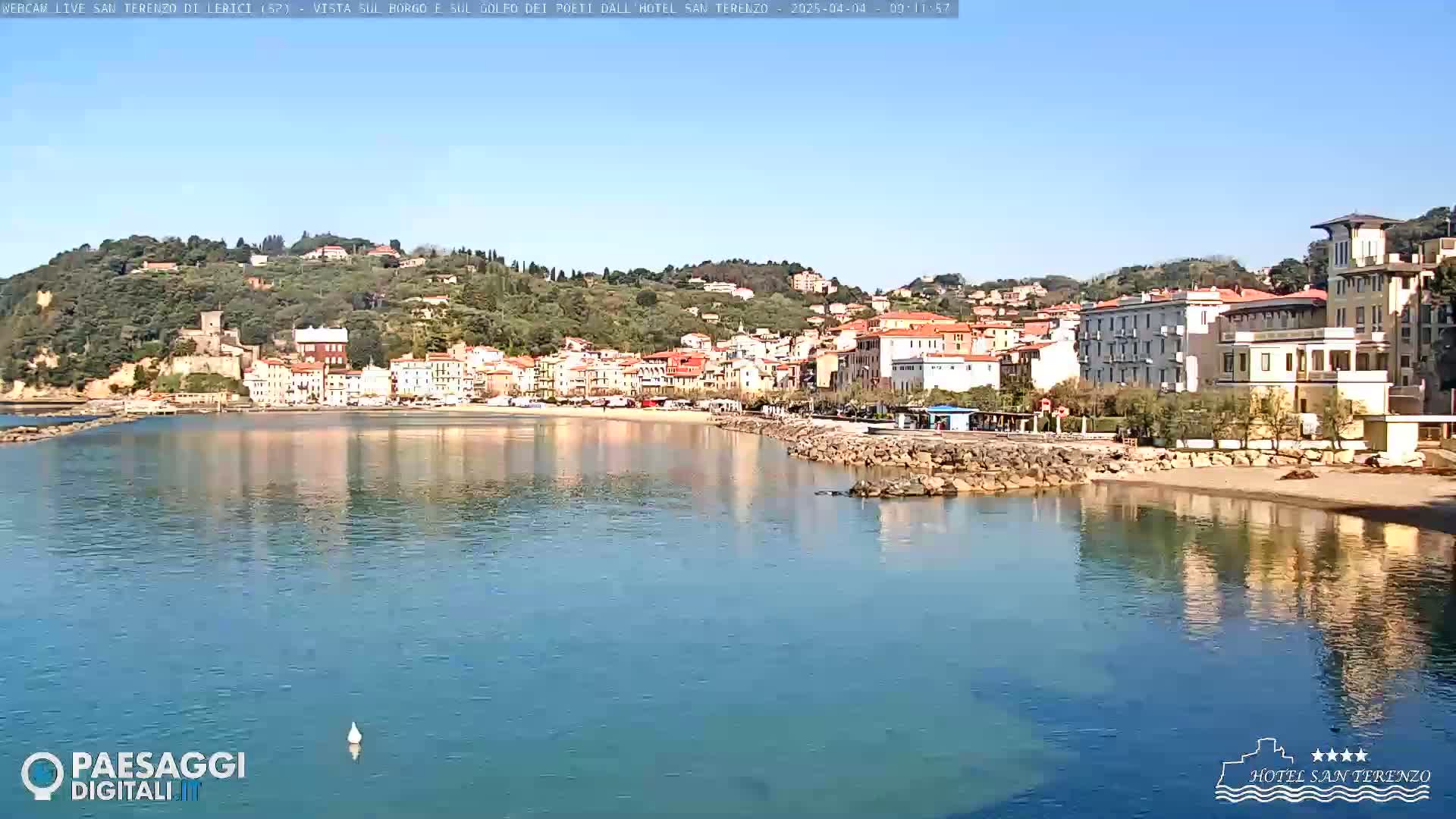 A coastal town with buildings on a hillside overlooking a calm, clear bay on a sunny day.