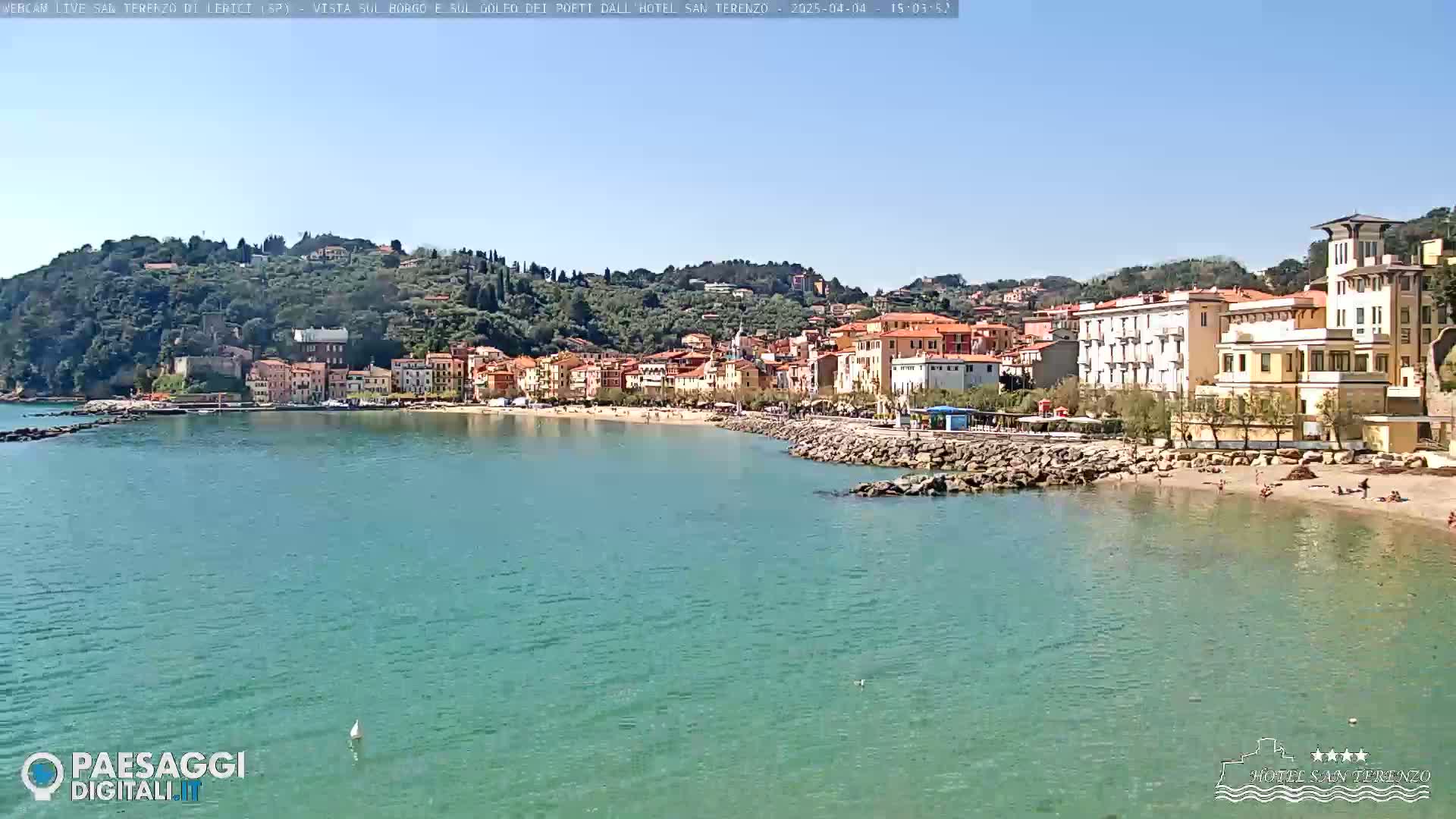 A picturesque coastal town, nestled against a verdant hillside, sits on a calm, clear, teal sea under a bright, sunny sky.