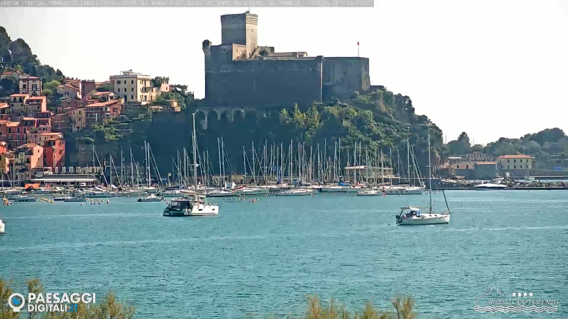 A stone castle sits atop a hill overlooking a harbor filled with sailboats on a sunny day.