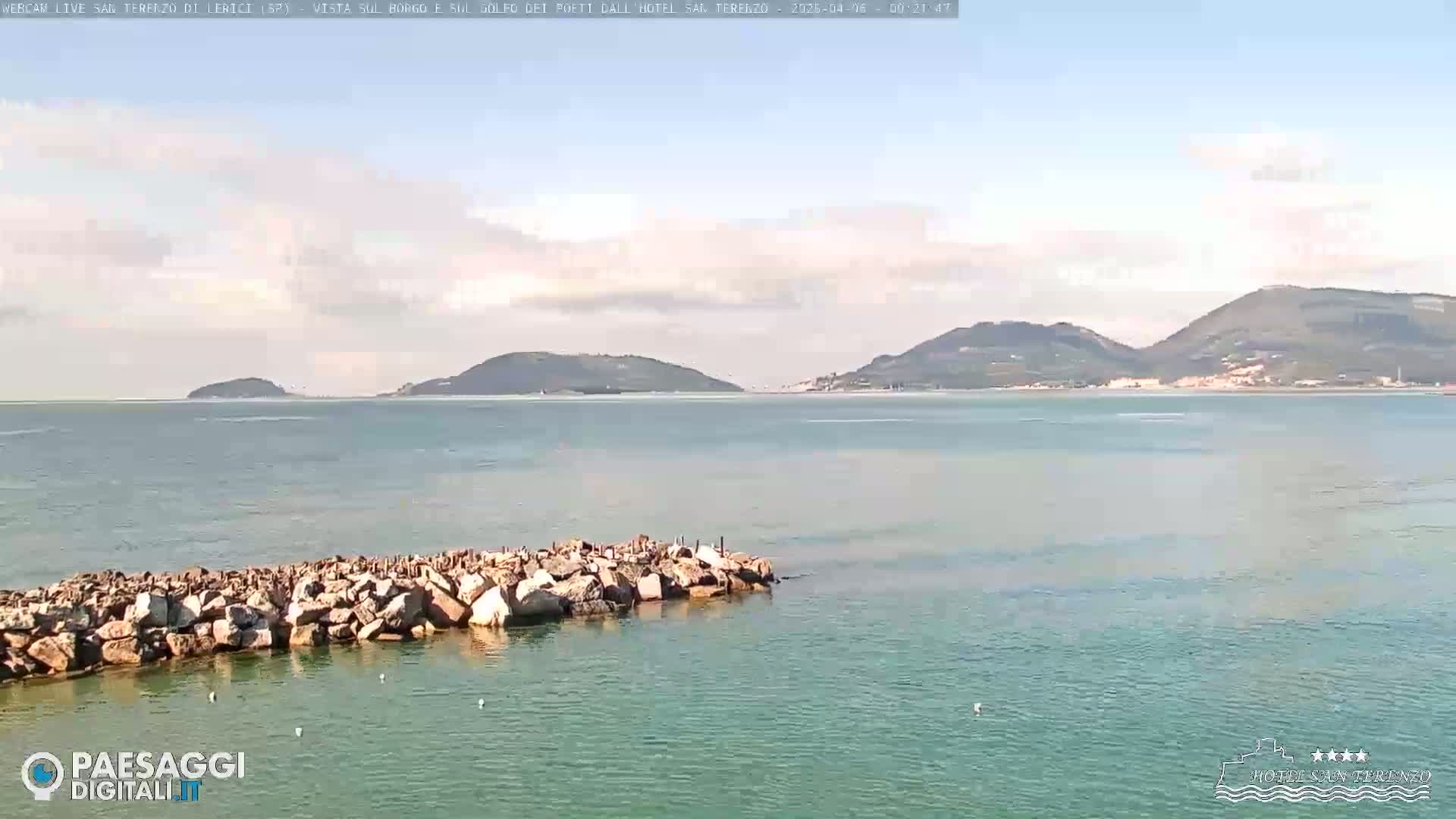A partly cloudy day shows a calm sea with a rock jetty in the foreground and distant hills across a bay.