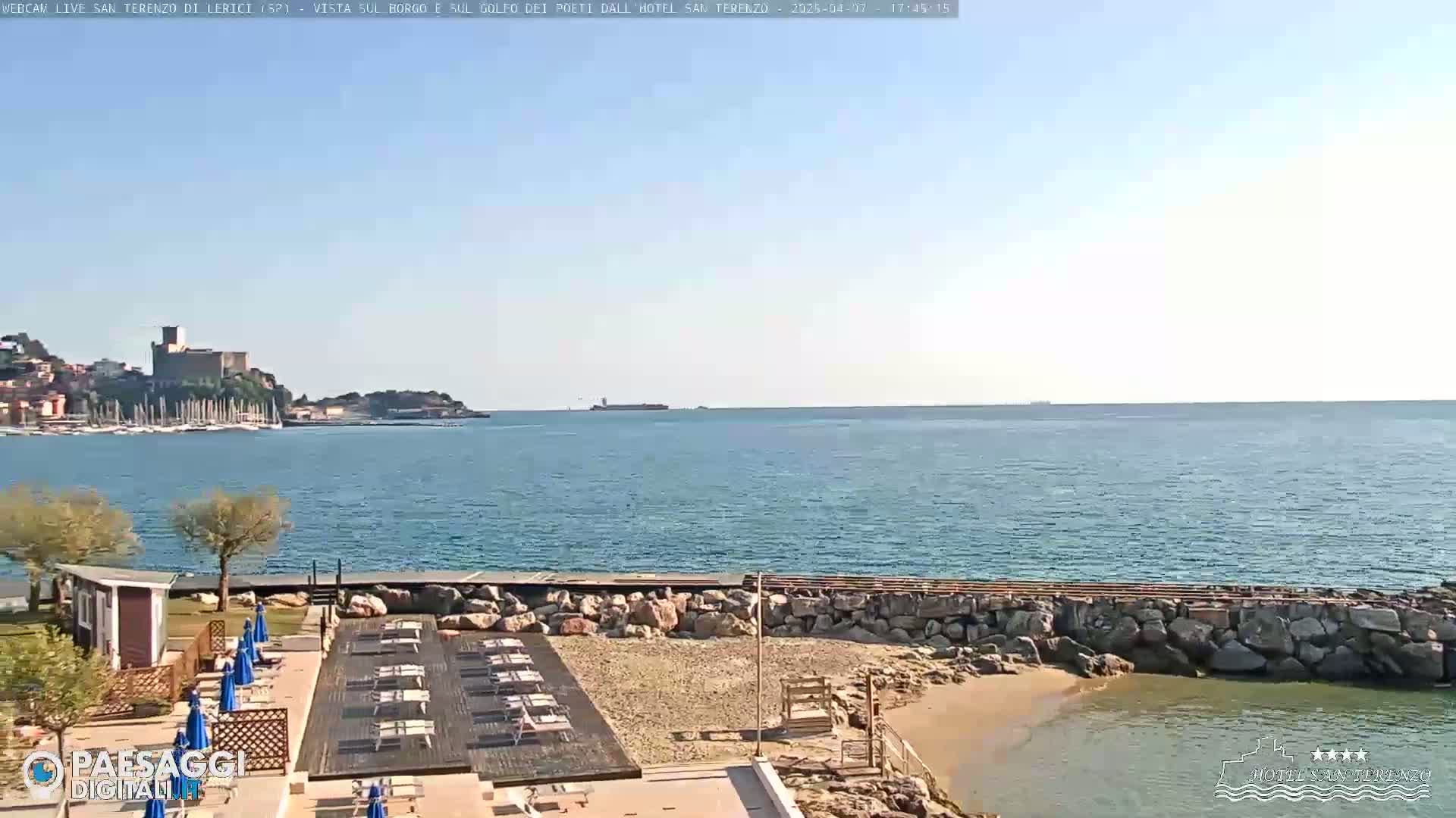 A seaside resort with lounge chairs and umbrellas overlooks a calm sea with a distant town and castle under a clear sky.