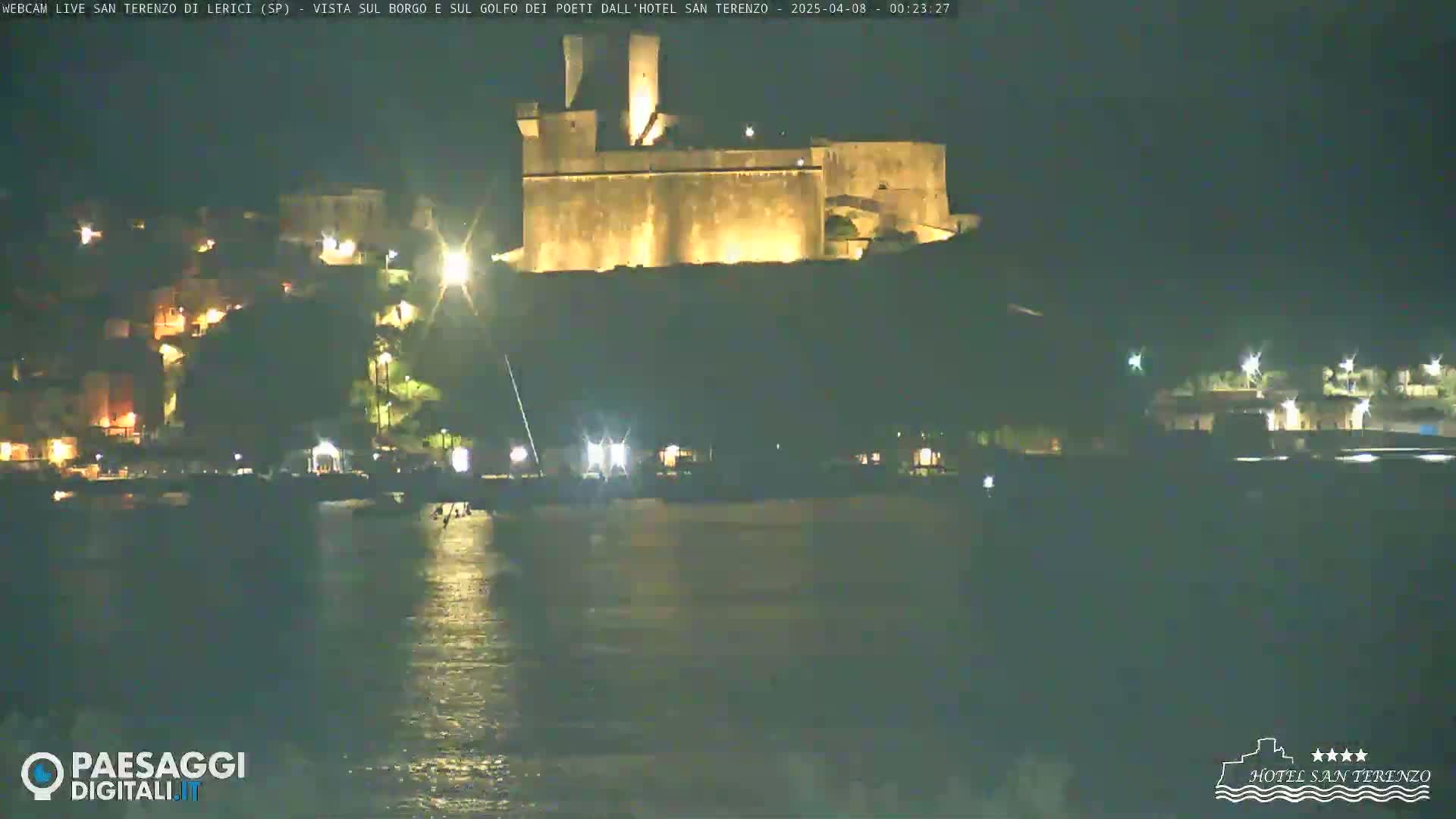 A nighttime view across calm water shows a hilltop village with a large illuminated stone structure, possibly a castle or fortress.