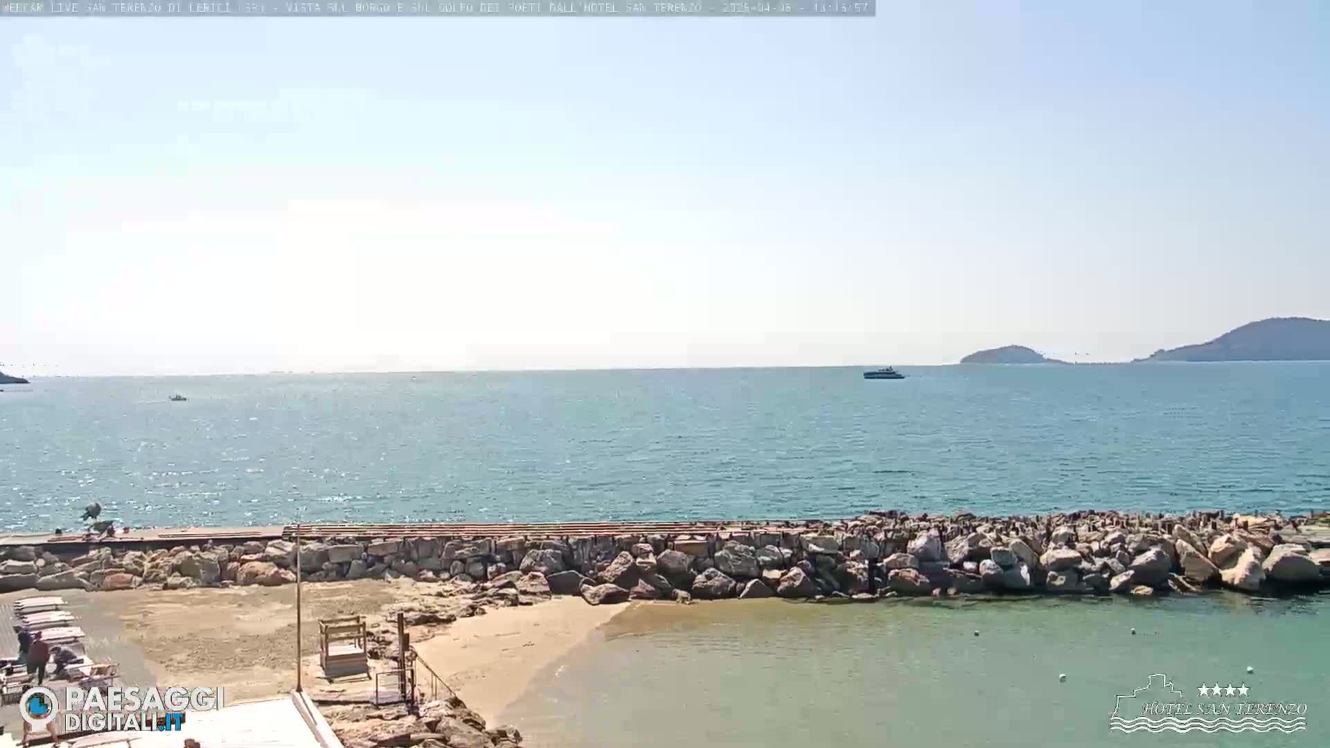 A rocky breakwater protects a small sandy beach area, with a calm sea extending to distant hills under a clear, sunny sky.