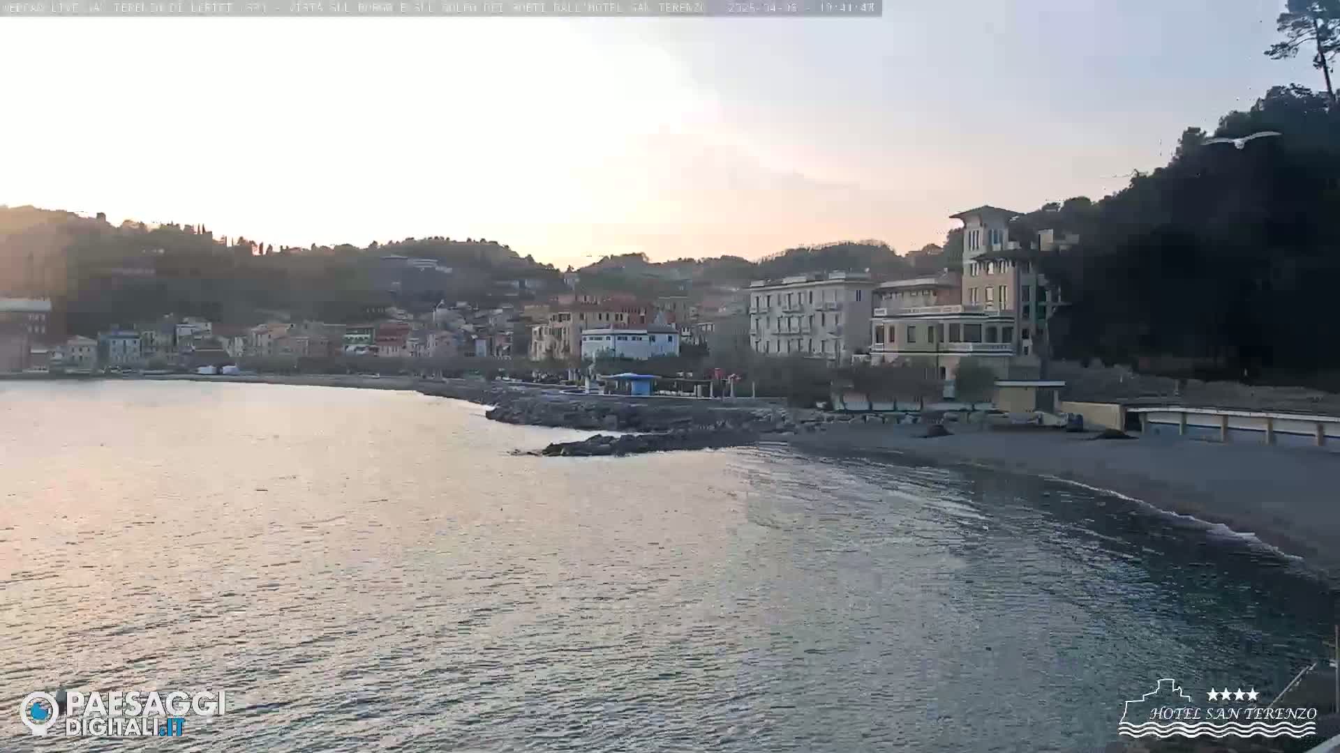 A coastal town with multi-story buildings sits on a calm, dark-water bay under a twilight sky.
