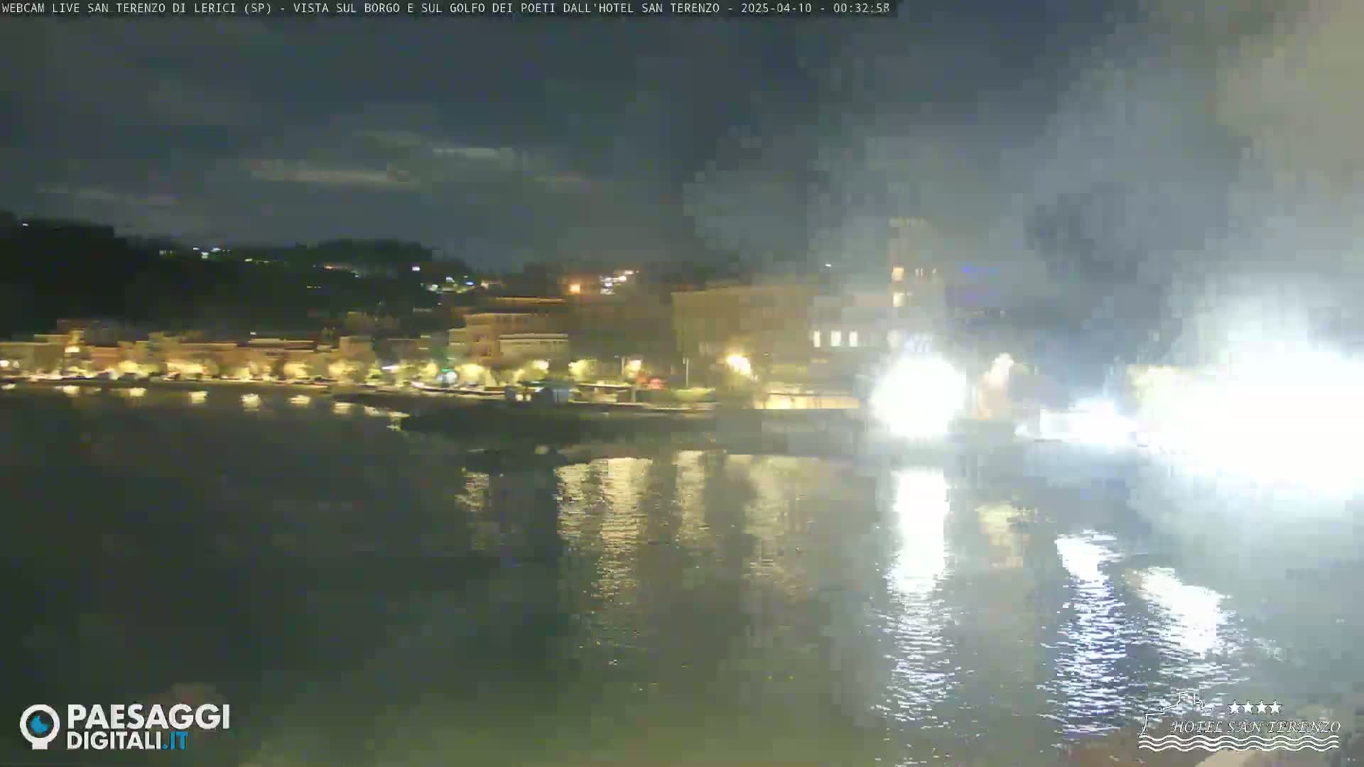 A nighttime view of a waterfront town with illuminated buildings, reflecting lights on the water, and a fireworks display over the water.