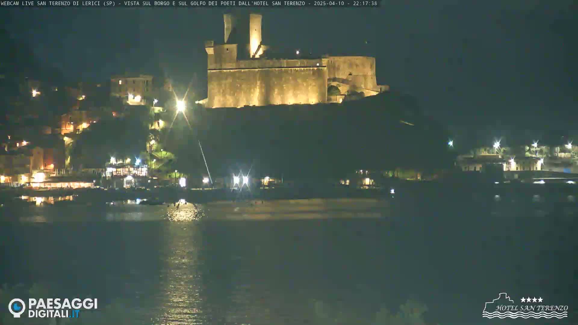 A nighttime view of a coastal town with a lit-up castle perched on a hill overlooking a dark body of water.