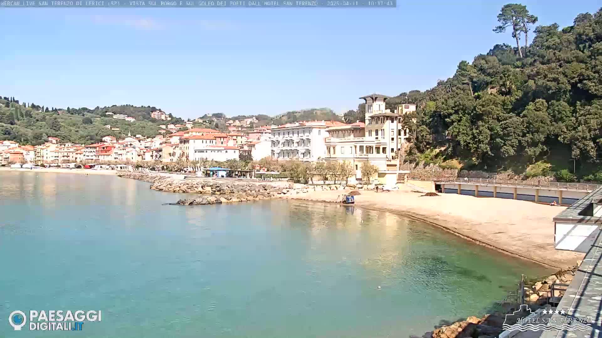 A beach with light-colored sand and clear turquoise water is situated below a seaside town nestled against a green hillside under a sunny sky.
