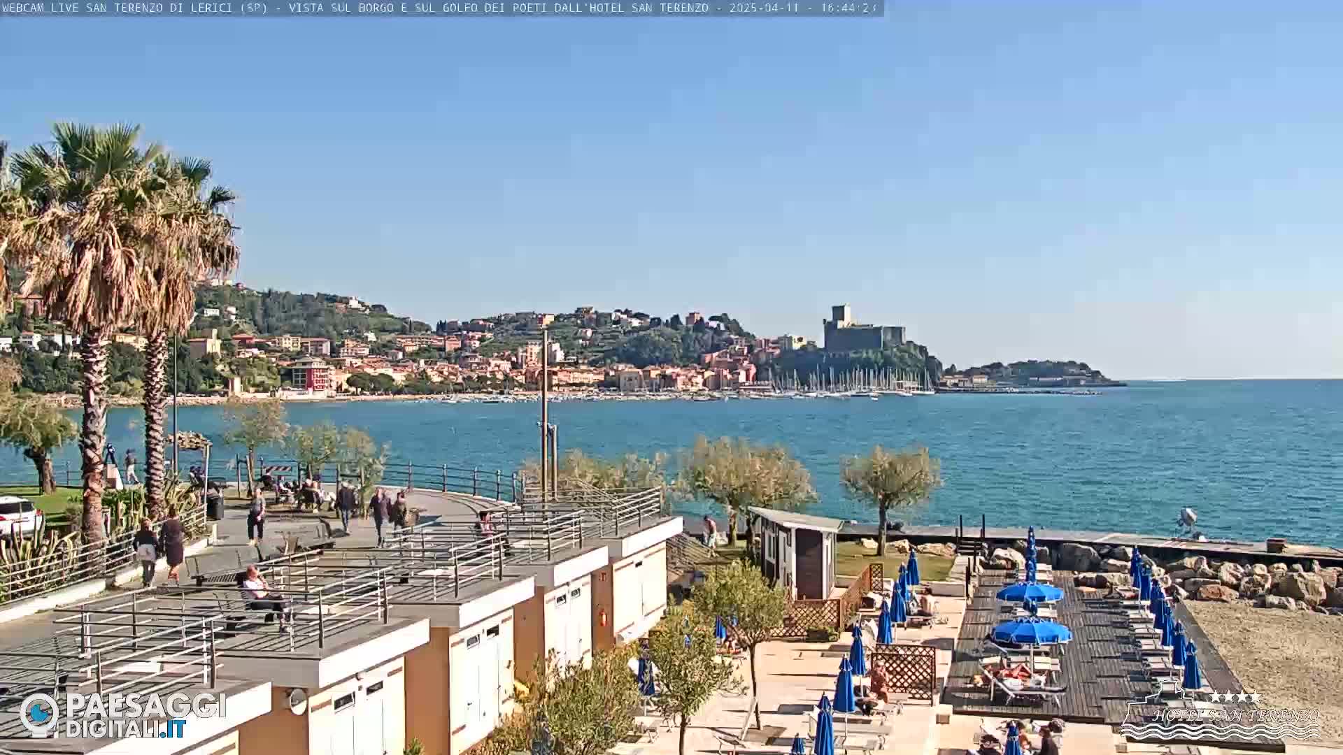 A sunny seaside town with a castle in the distance is viewed from a beachside promenade with people walking and relaxing on lounge chairs under umbrellas.