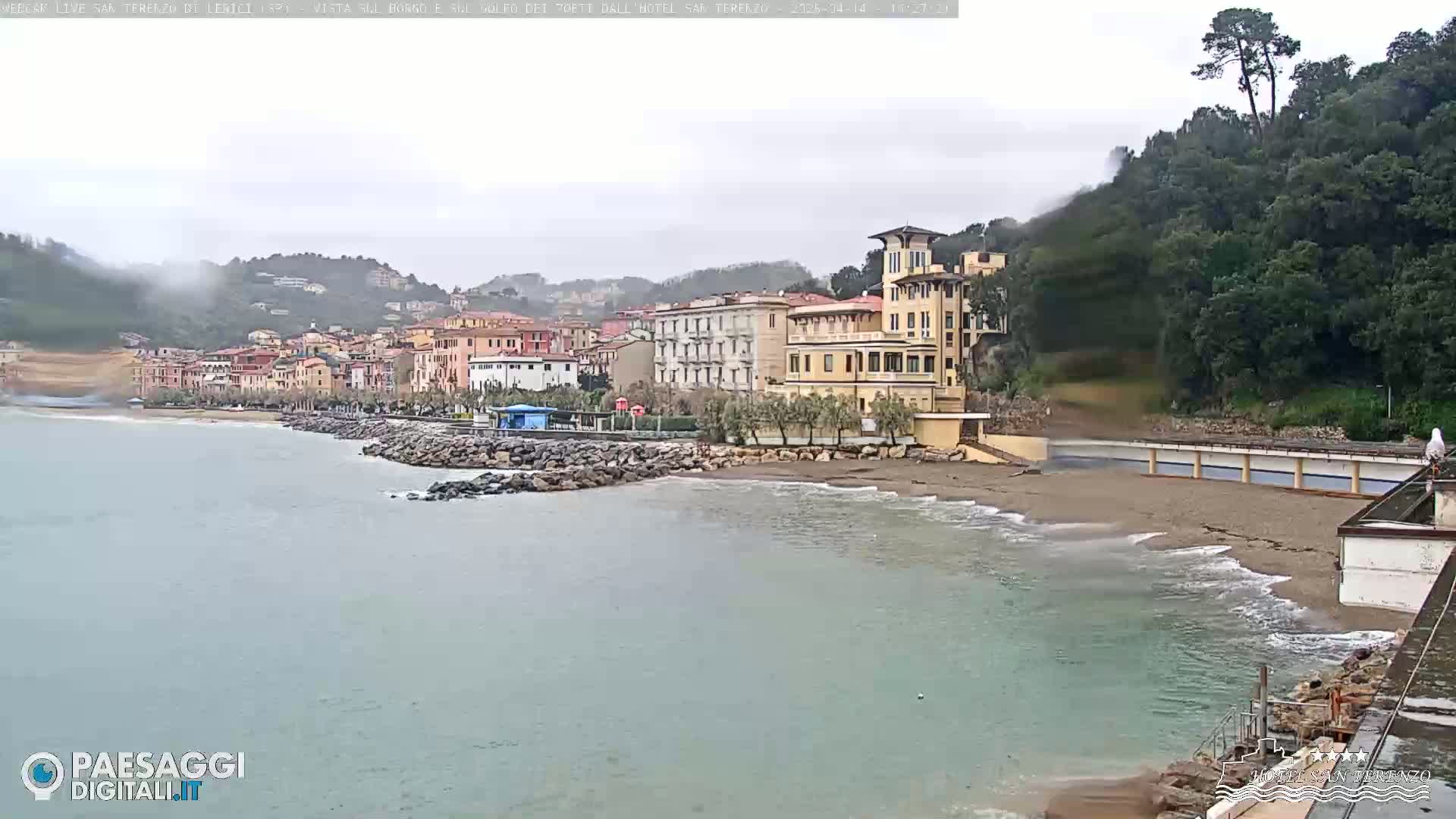 A seaside town with multi-story buildings sits on a gently sloping coastline under an overcast sky, with small waves lapping a sandy beach.