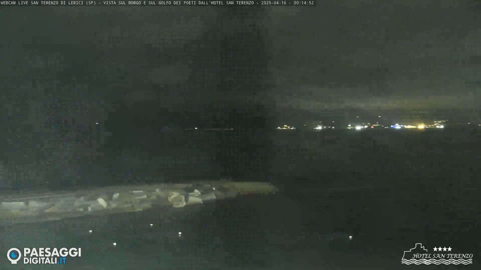 A nighttime, long shot overlooking a dark body of water with faintly visible lights across the horizon under a cloudy sky.