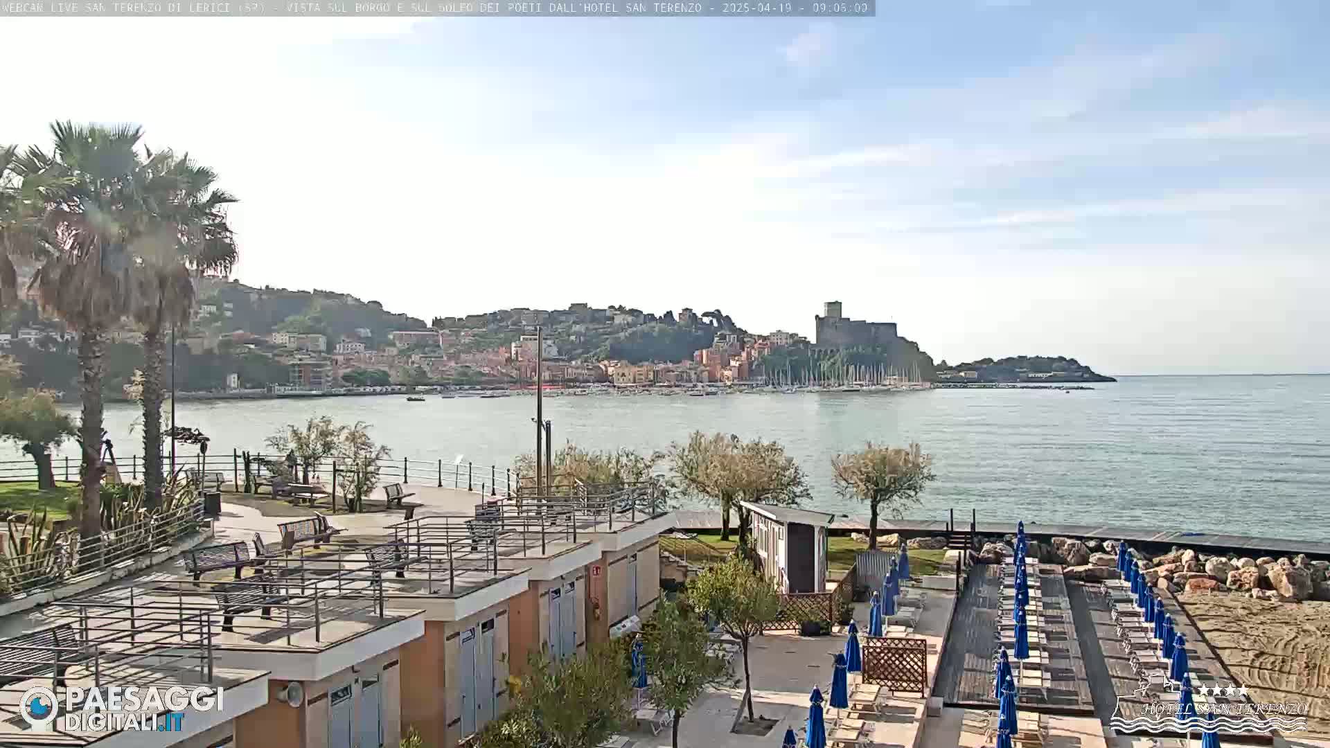 A seaside town with a castle on a hill is viewed from a beach area with lounge chairs and umbrellas under a mostly clear sky.