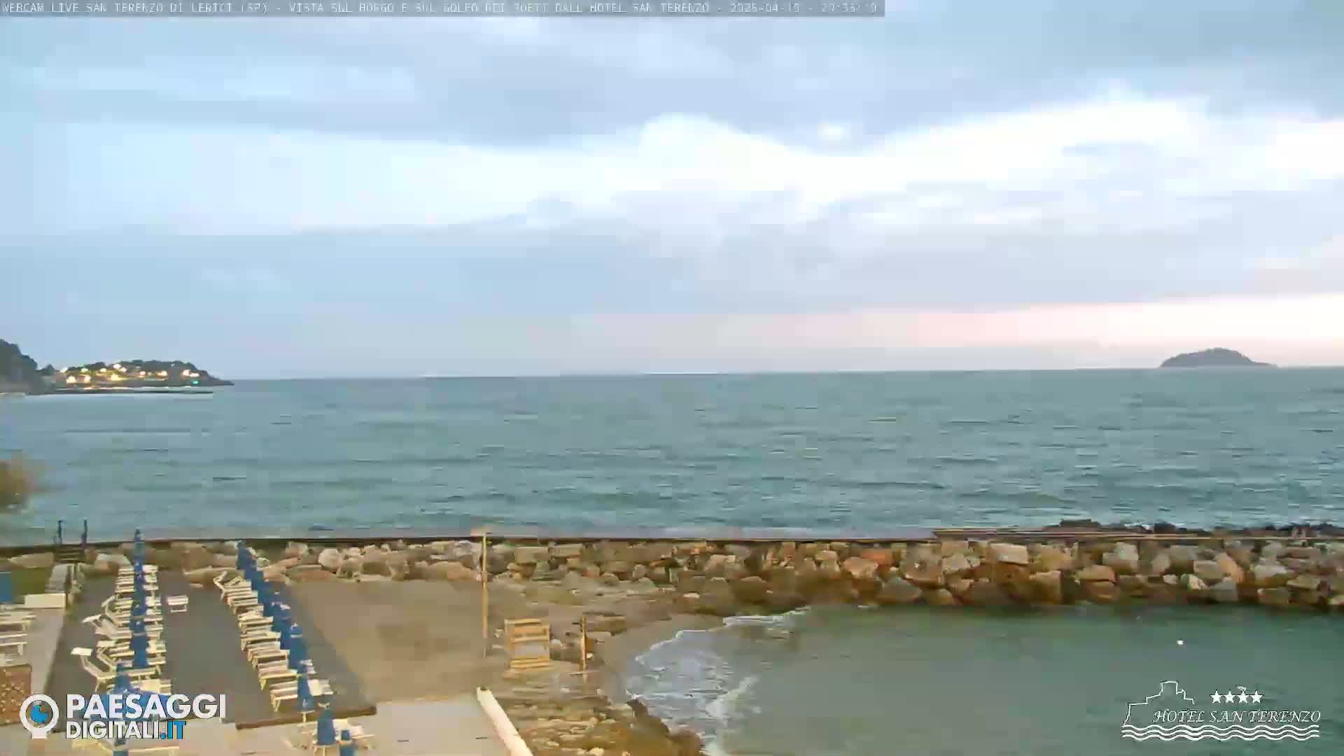 A partly cloudy day shows a calm sea with a rocky shoreline and rows of beach chairs.