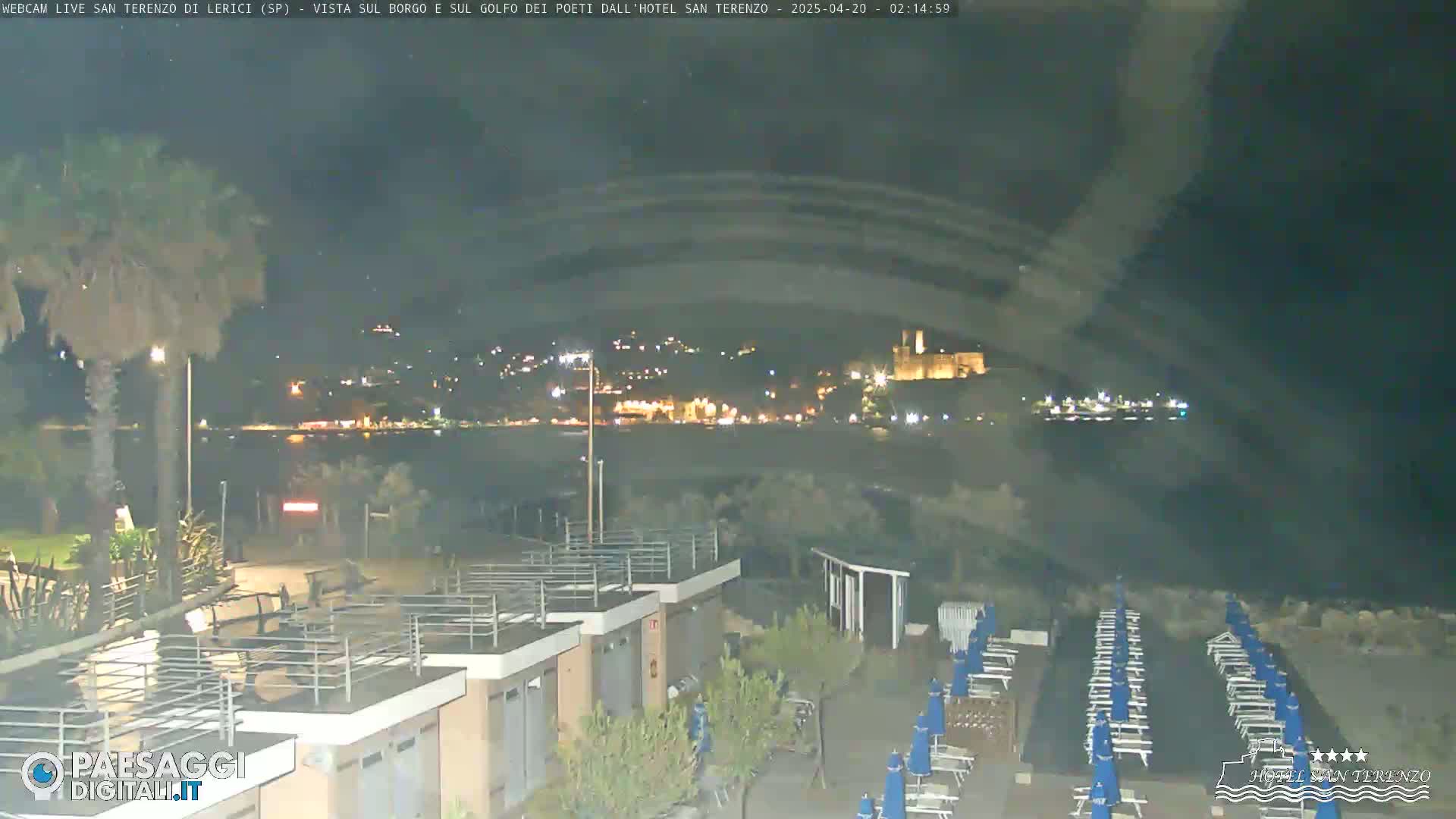A nighttime view from a beachfront property shows rows of beach chairs and umbrellas, with a distant, illuminated town and calm water visible beyond.