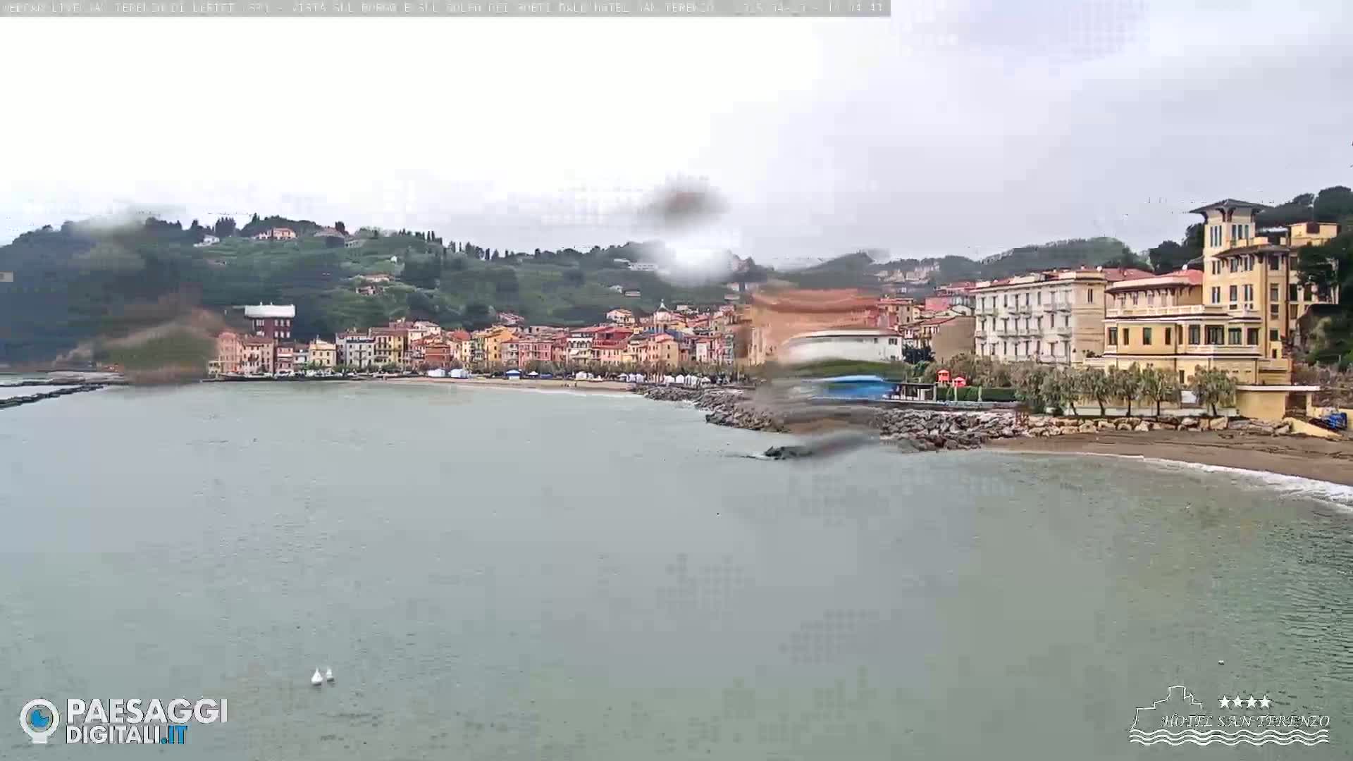 A coastal town with buildings along a sandy beach is seen on an overcast day, with a calm sea in the foreground.