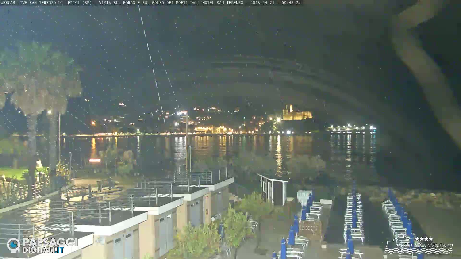 A nighttime, rain-swept view shows a calm body of water reflecting the lights of a town across the shore, with beach chairs and umbrellas set up in the foreground.