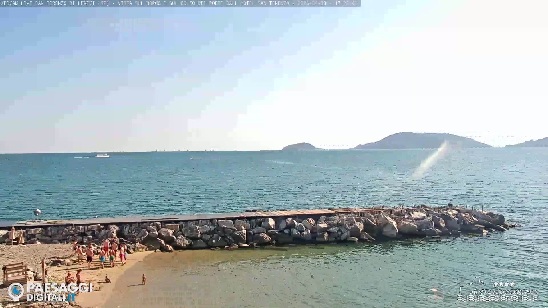 A rocky breakwater separates a small sandy beach with people on it from a calm sea under a mostly clear sky, with distant hills visible on the horizon.