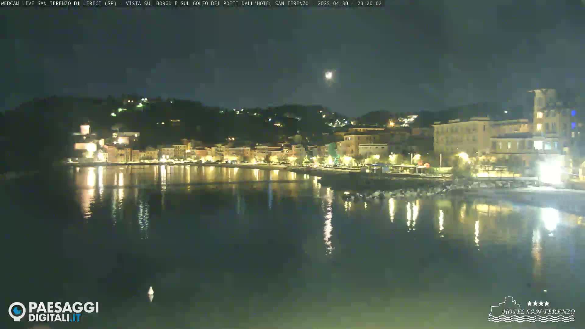 A nighttime view of a coastal town illuminated with lights, reflected in the calm water under a clear night sky with a visible moon.