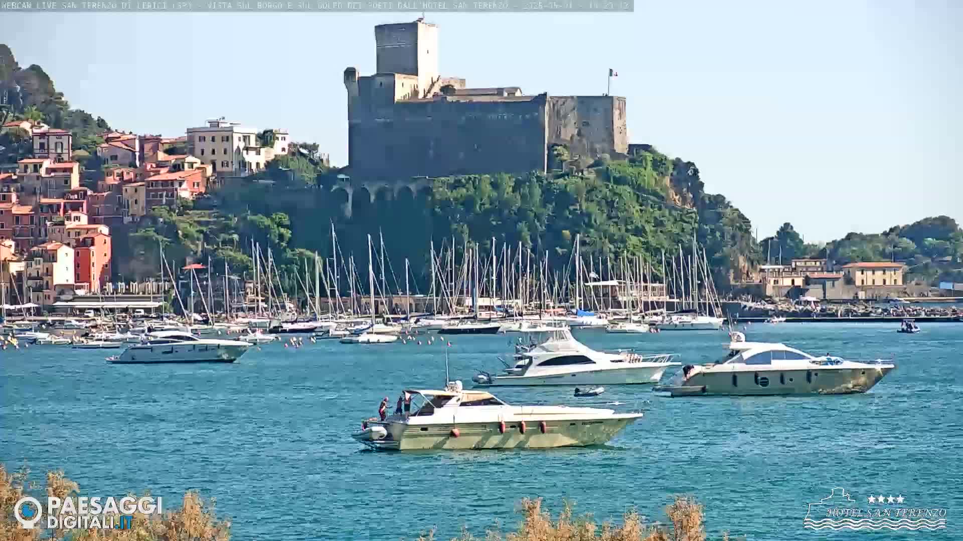 A stone castle sits atop a hill overlooking a harbor filled with sailboats and motor yachts on a sunny day.